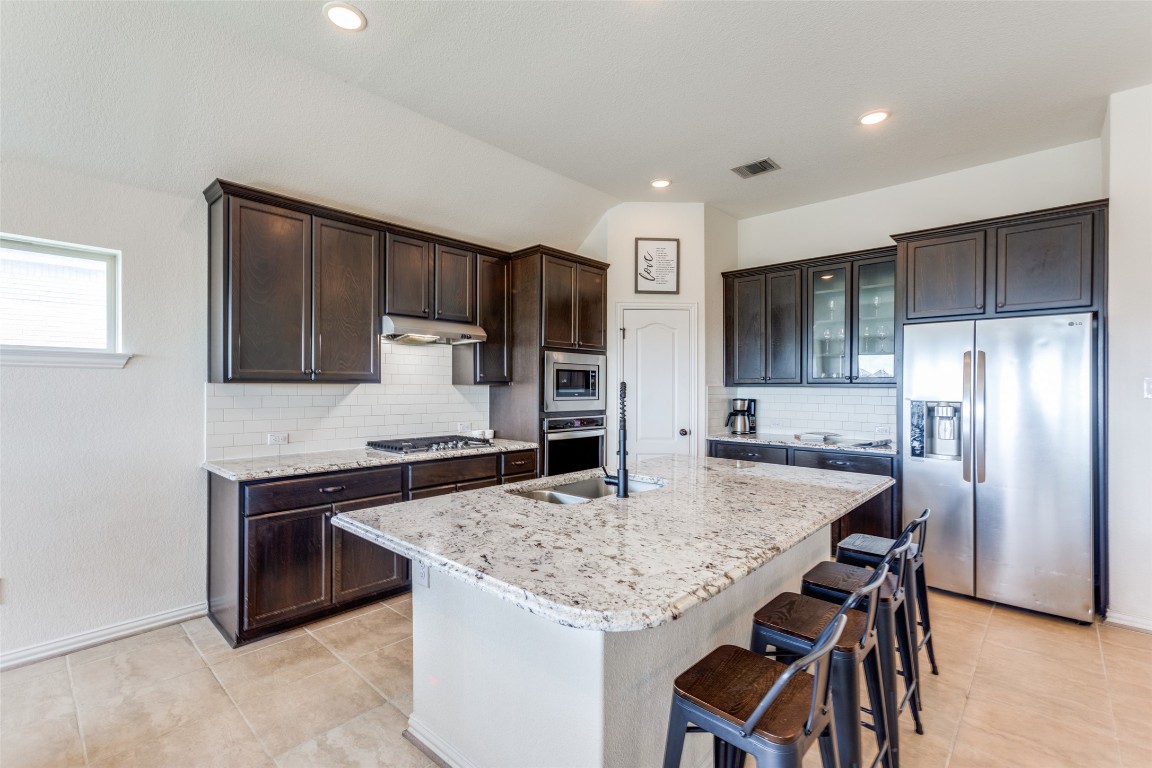874 Spinnaker Loop Kyle, TX 78640 - Photo 7 of 30 Kitchen with appliances with stainless steel finishes, light stone counters, a kitchen island with sink, dark brown cabinets, and a kitchen breakfast bar