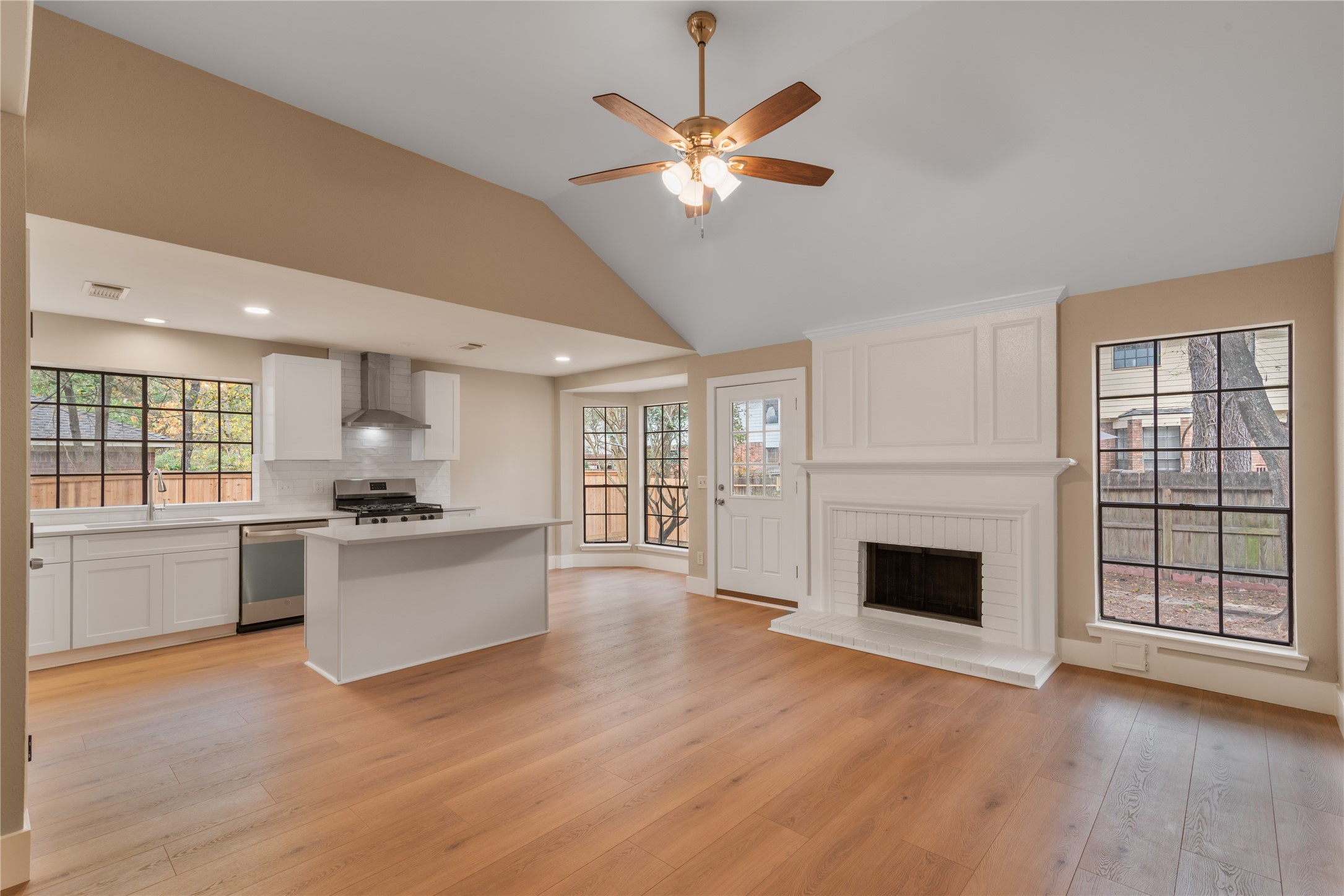 10 White Fawn Drive Spring, TX 77381 - Photo 7 of 25 This photo showcases a bright, open-concept living area featuring a modern kitchen with white cabinets, a central island, and stainless steel appliances. The space includes a cozy fireplace, large windows providing ample natural light, and hardwood flooring throughout. A ceiling fan enhances the airy, welcoming atmosphere.