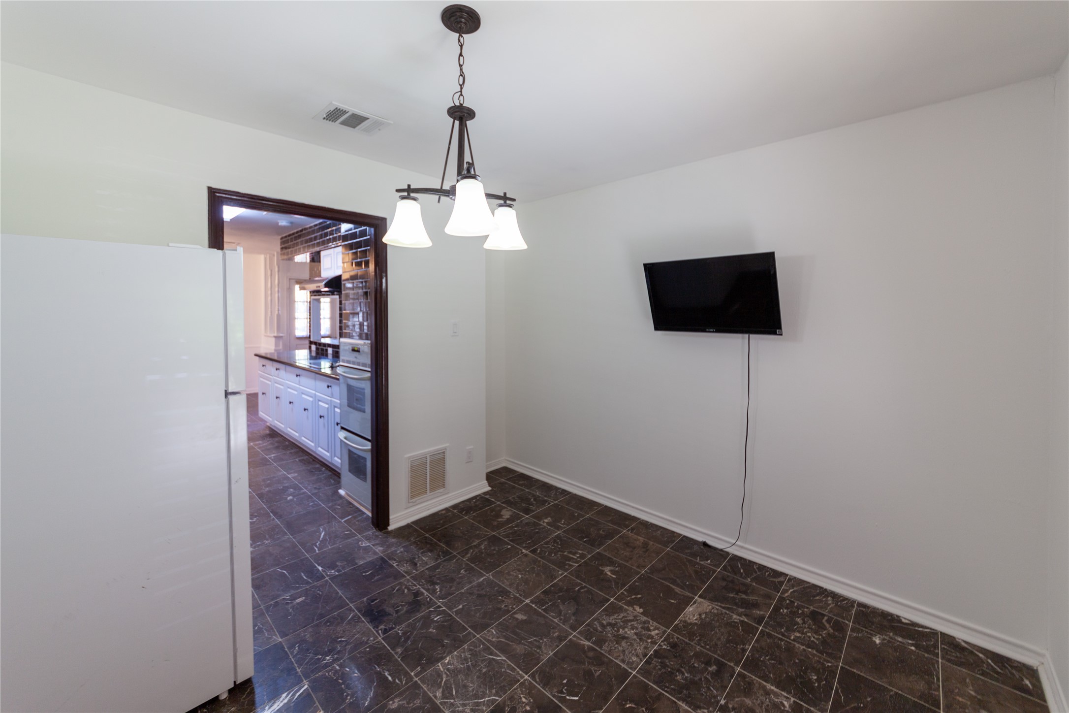 8826 Roos Road Houston, TX 77036 - Photo 11 of 42 Bright dining area with dark tiled floors, a wall-mounted TV, and a modern light fixture. It opens into a kitchen with white cabinetry and stainless steel appliances, offering a clean and functional space.