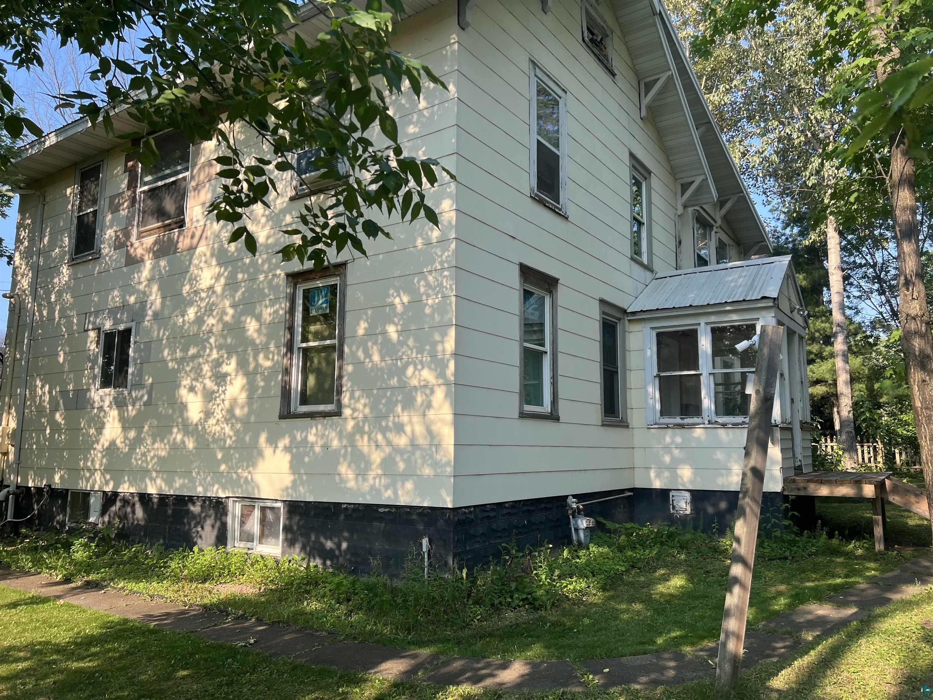 107 6th Street South Virginia, MN 55792 - Photo 2 of 7 Light-colored siding exterior featuring multiple windows, a covered porch with a metal roof, and a partial wooden deck