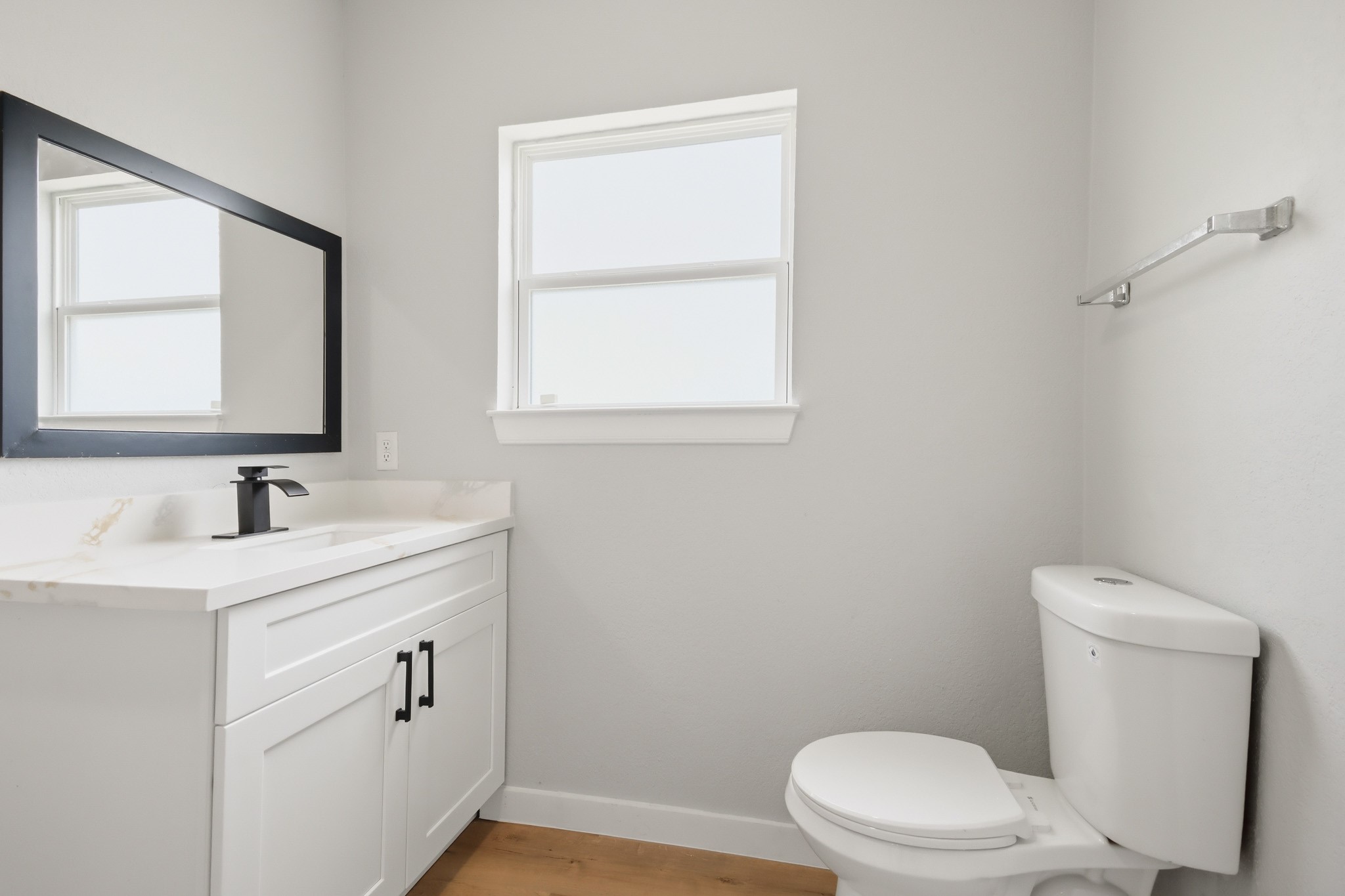 10430 Wiggins Street Houston, TX 77029 - Photo 17 of 27 Primary bathroom features a clean, modern design with a white vanity, sleek black fixtures, and a large mirror. It has a bright window, light gray walls, and wooden flooring, creating a fresh and inviting atmosphere.