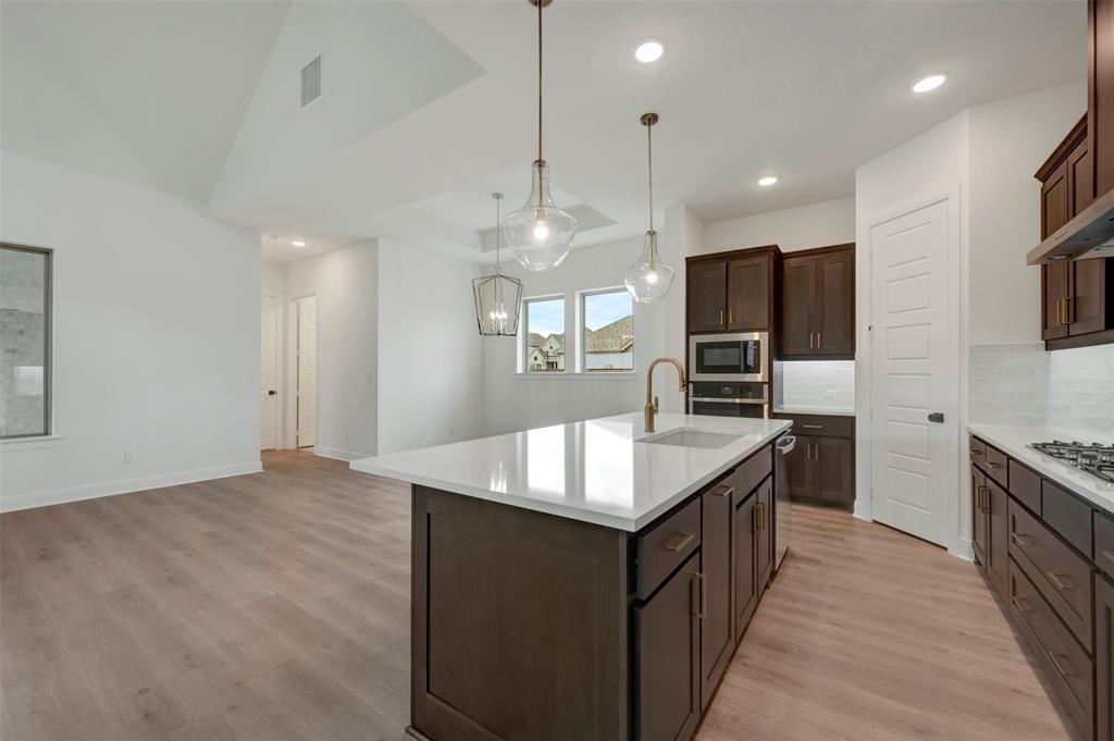 1908 Cola Drive Van Alstyne, TX 75495 - Photo 12 of 29 Open-concept kitchen featuring a central island with a white countertop and integrated sink, pendant lighting, dark wood cabinetry with gold hardware, stainless steel appliances, and wood-finish flooring