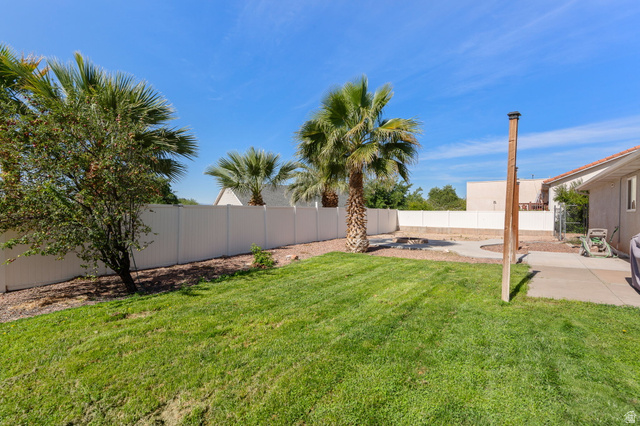 664 East Springhill Drive Washington, UT 84780 - Photo 18 of 21 Spacious backyard featuring a large grass area, mature palm trees, white perimeter fencing, concrete patio, and a gravel-filled area with a circular fire pit
