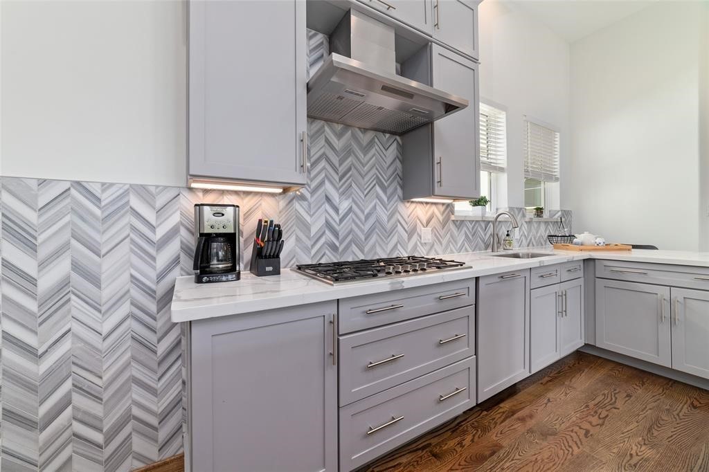 710 Delano Street, Unit A Houston, TX 77003 - Photo 16 of 32 The modern kitchen features sleek gray cabinetry and a stylish chevron-patterned backsplash. It includes a gas stove, stainless steel range hood, and ample counter space with a built-in sink, all set against warm hardwood floors.