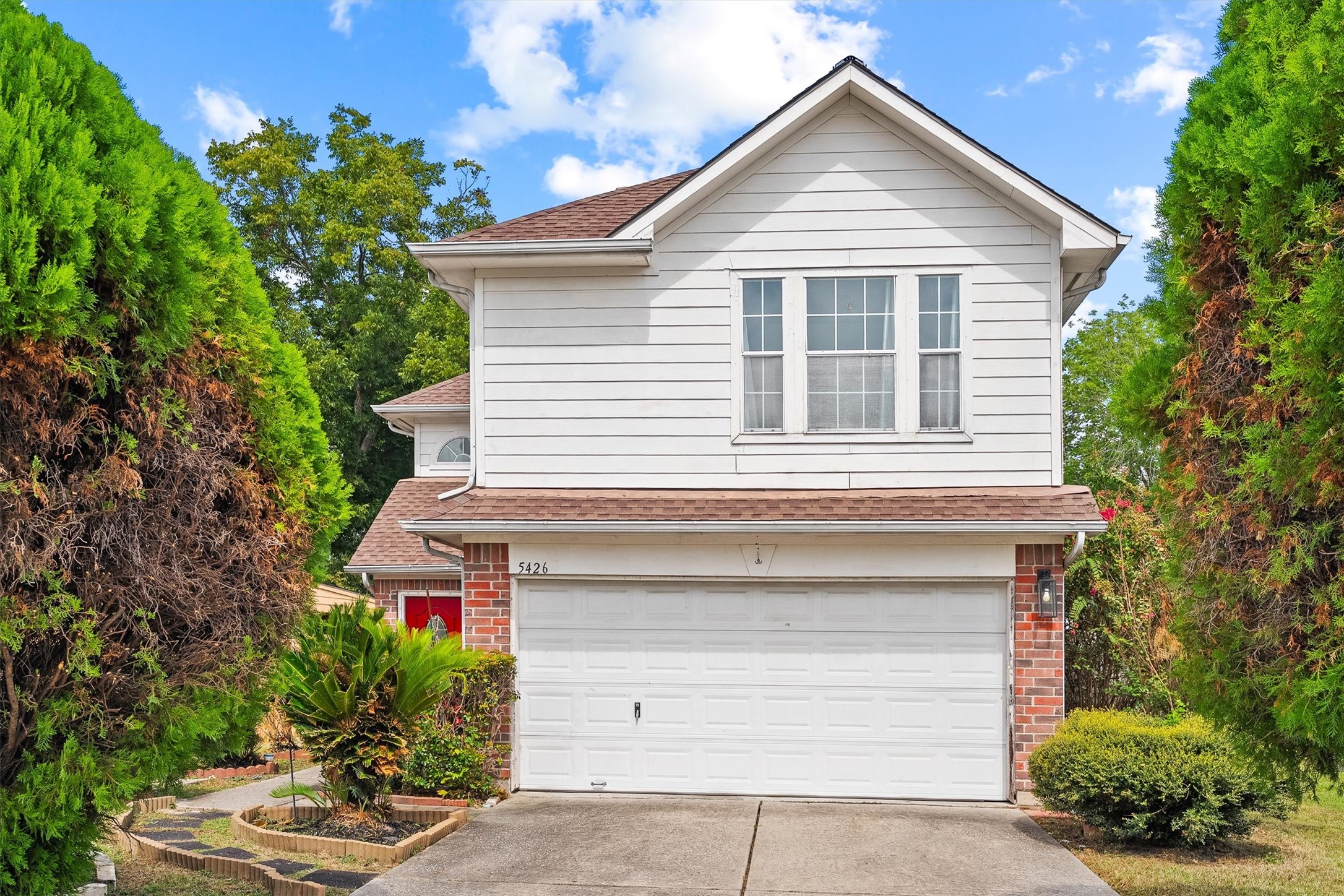 Attractive two-story home with a classic white exterior and brick accents, featuring a spacious two-car garage and lush greenery framing the entrance.