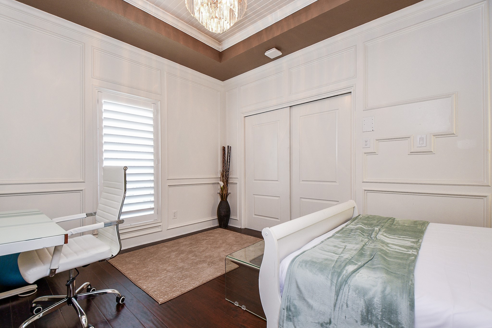 263 Wood Loop Street Houston, TX 77015 - Photo 44 of 45 This room features elegant wainscoting, a coffered ceiling with a modern chandelier, and rich hardwood floors. It includes a cozy nook with a bed, a sleek glass desk with a white office chair, and a closet with sliding doors, offering a stylish and functional space.