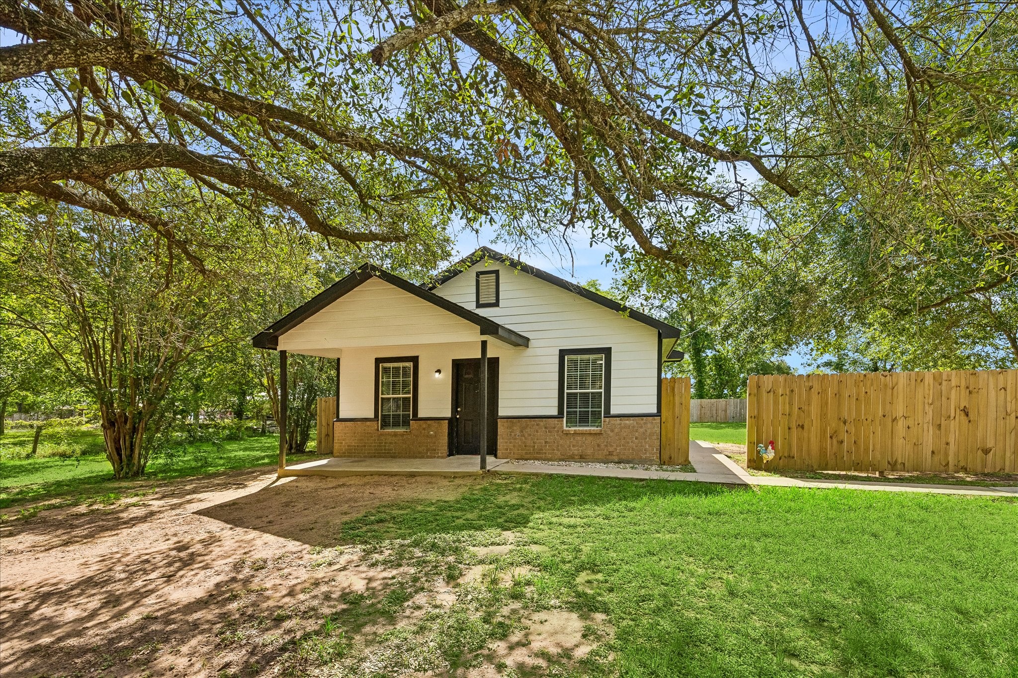 Charming single-story home with a welcoming porch, nestled under mature trees. Features a combination of brick and siding, a fenced backyard, and a spacious lawn.