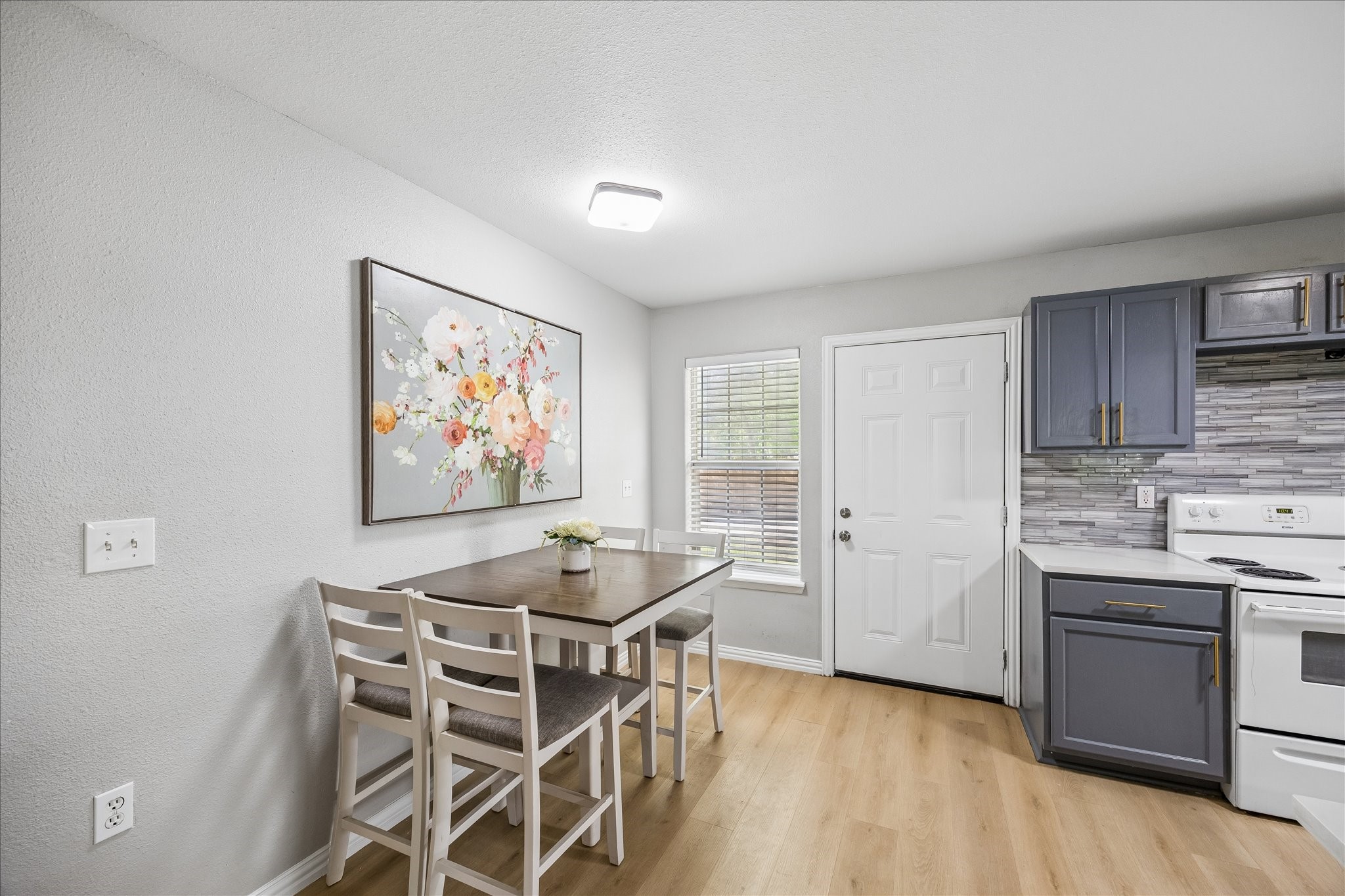 539 Anderson Street Sealy, TX 77474 - Photo 12 of 19 Cozy kitchen-dining area with light wood flooring and soft gray walls. Modern cabinetry in a muted blue-gray tone. The space is well-lit with natural light from a window and a sleek ceiling light fixture.