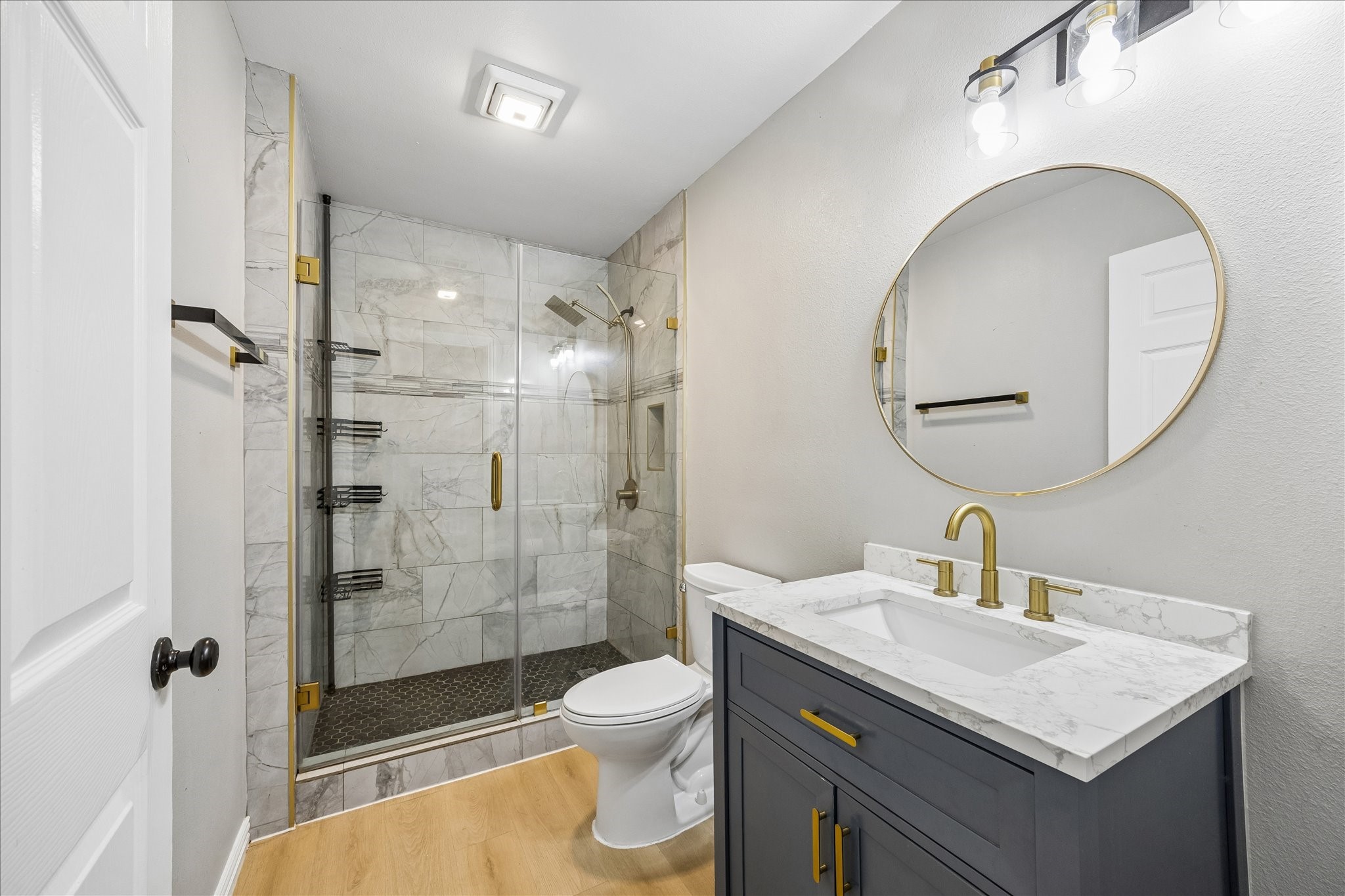 539 Anderson Street Sealy, TX 77474 - Photo 13 of 19 This bathroom features a modern design with a sleek glass-enclosed shower, marble-like tiles, and gold fixtures. It includes a stylish vanity with a round mirror and ample lighting, complemented by a light wood floor.