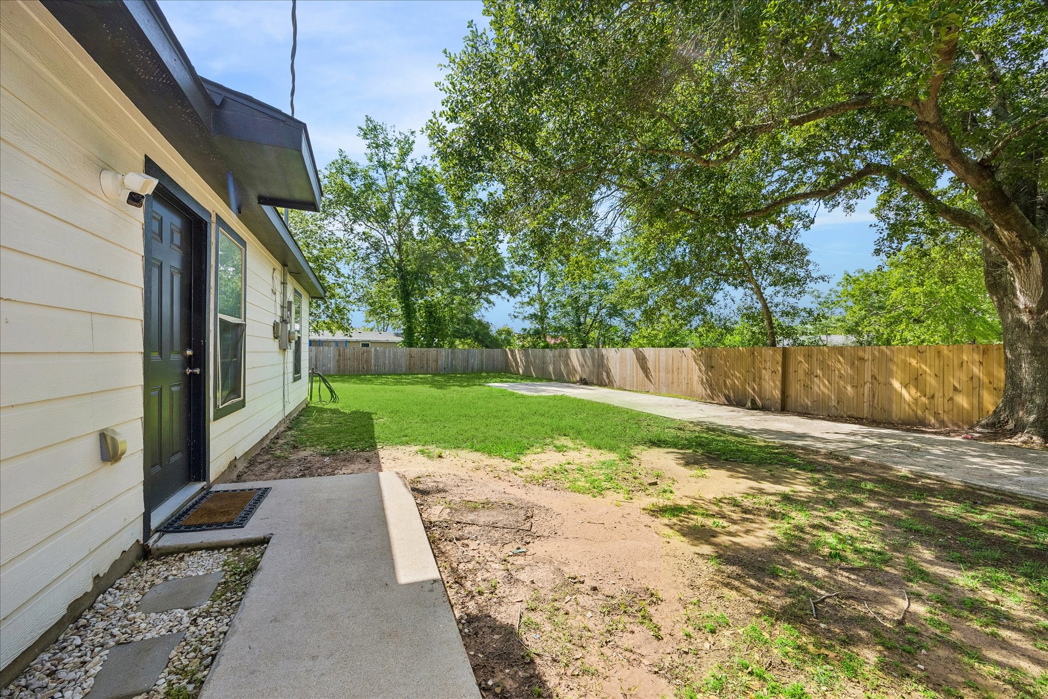 539 Anderson Street Sealy, TX 77474 - Photo 17 of 19 Spacious backyard with a well-maintained lawn and a large tree providing shade. The house exterior features light siding and a black door, with a concrete path leading to the yard. A wooden fence offers privacy.