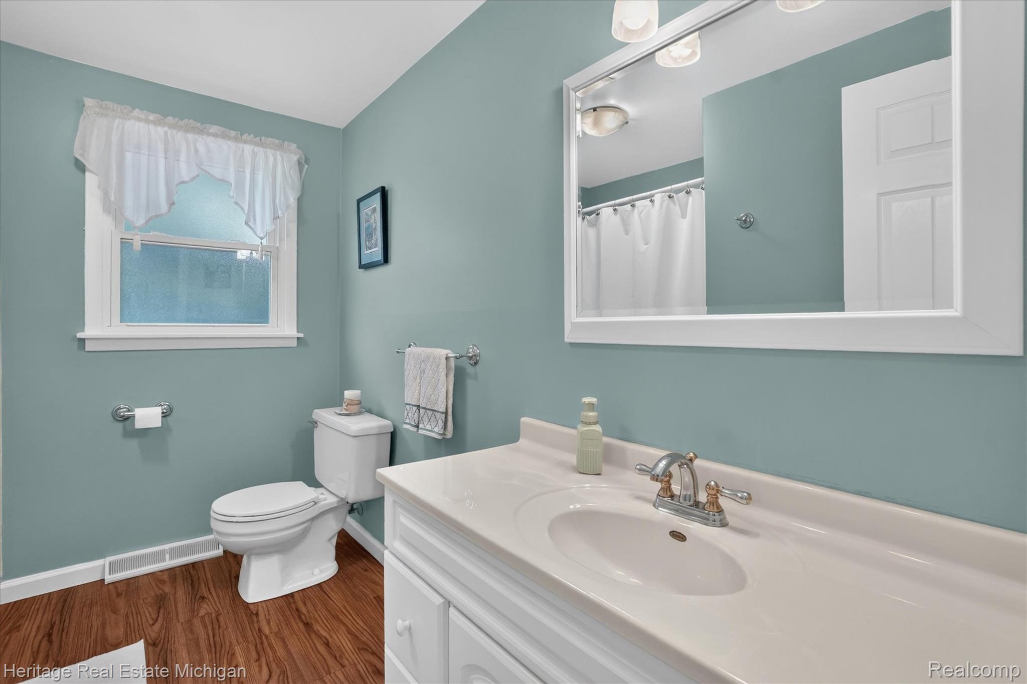 14038 North Hogan Road Linden, MI 48451 - Photo 29 of 53 Bathroom featuring a vanity with an integrated sink and brushed nickel faucet, framed mirror, wall-mounted light fixture, white toilet, and wood-finish flooring