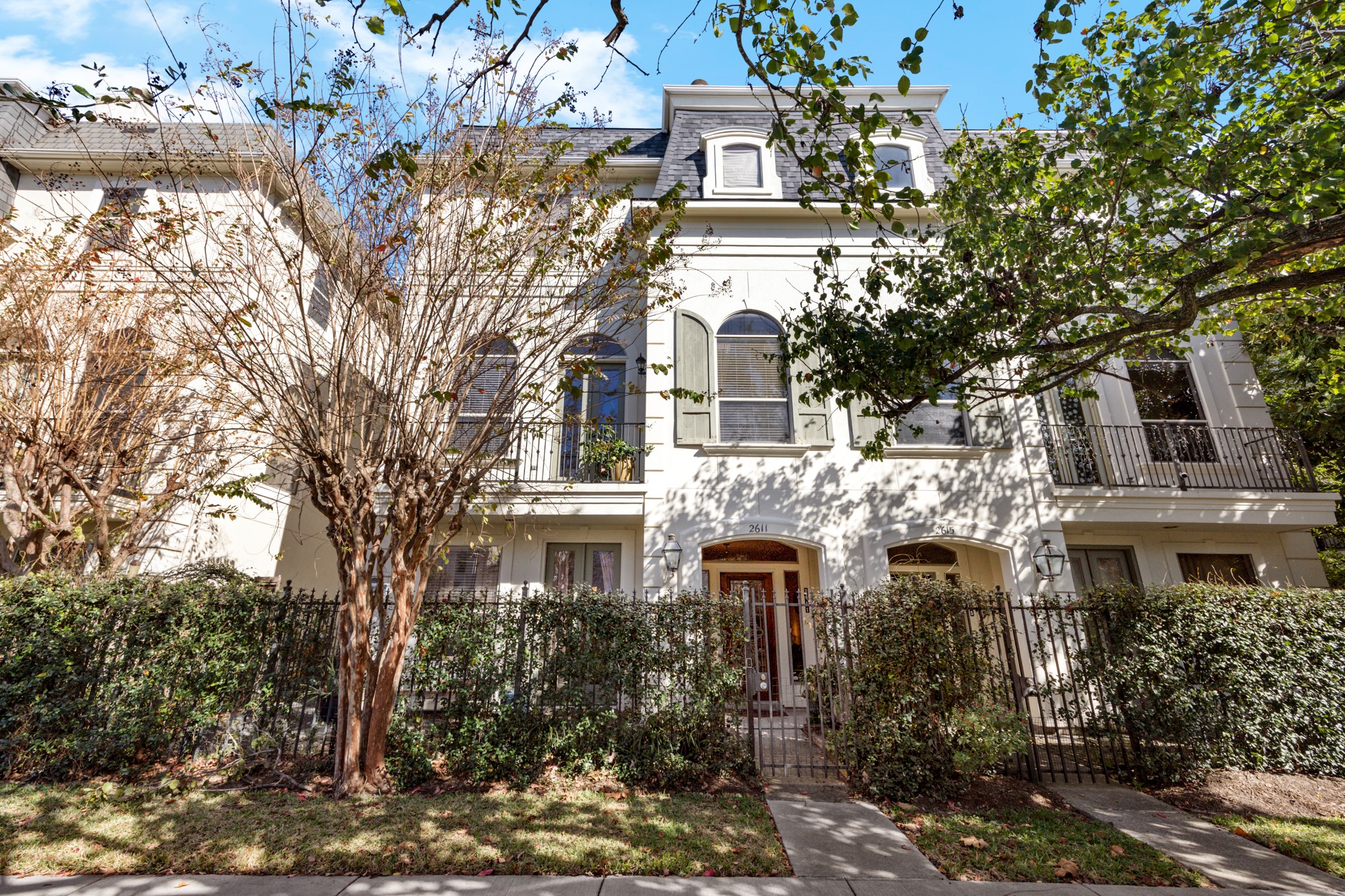 2611 Commonwealth Street Houston, TX 77006 - Photo 2 of 31 Beautiful three-story townhome with a refined, classic façade. The exterior features a light, elegant finish with symmetrical architectural details, arched windows, and decorative rooflines that give it a timeless feel. A wrought-iron gated entry leads to a private front door, framed by manicured hedges and mature trees that add both privacy and curb appeal. Upper levels include shuttered windows and small balcony railings, while the overall streetscape.