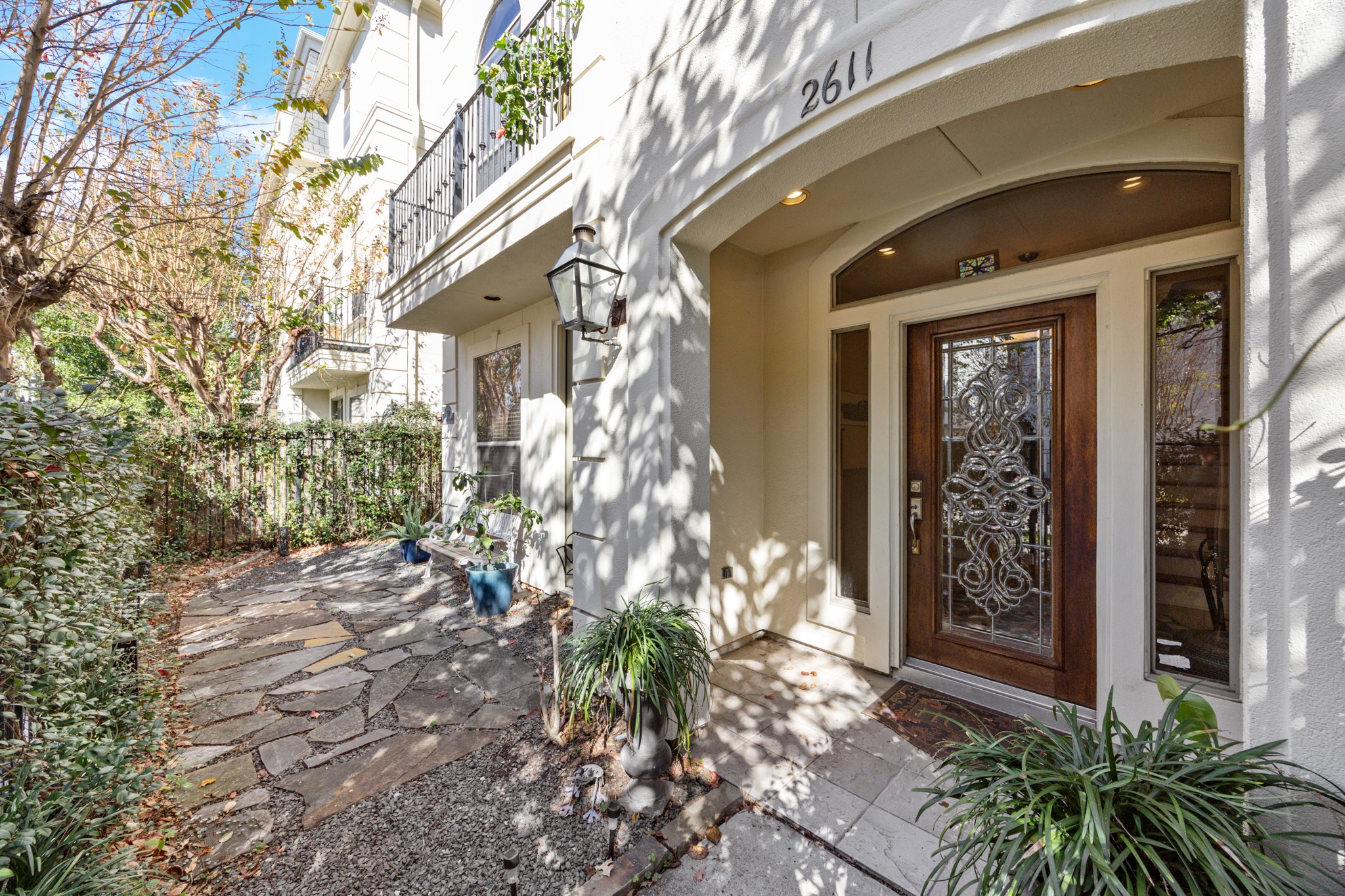 2611 Commonwealth Street Houston, TX 77006 - Photo 3 of 31 The inviting front entry of the townhome, where a private, gated walkway leads to a refined recessed doorway. A striking wood-and-glass front door is framed by an elegant arched entry and classic lantern-style lighting, creating a memorable first impression. The stone path winds through an easy-to-maintain courtyard with mature trees, potted plants, and curated landscaping—offering a serene, low-maintenance outdoor retreat that feels both private and welcoming