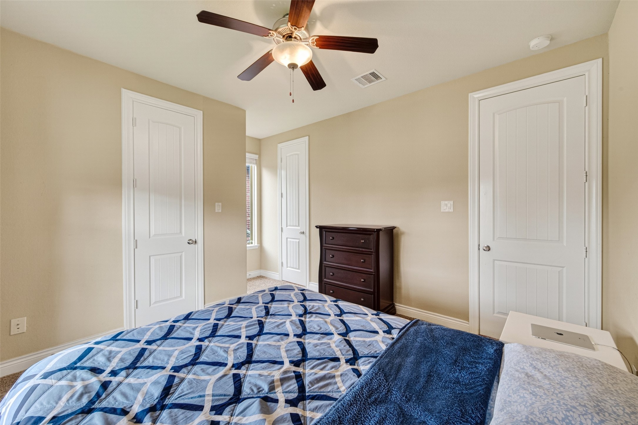 25611 Ellis Ridge Court Katy, TX 77494 - Photo 38 of 49 This cozy bedroom features neutral walls, a ceiling fan, and ample natural light. It includes two white doors, a dark wooden dresser, and a bed with a blue patterned comforter, creating a comfortable and inviting space.