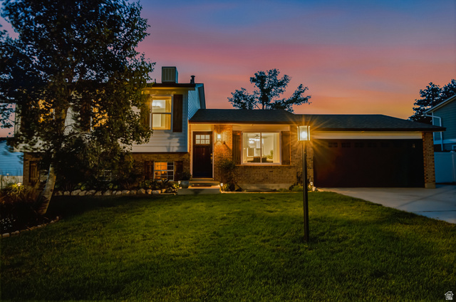12152 Cinnamon Ridge Road Sandy, UT 84094 - Photo 32 of 34 Brick Colonial home illuminated by landscape lighting, featuring a two-car attached garage, manicured lawn with mature trees, and a modern front entrance with illuminated architectural detailing
