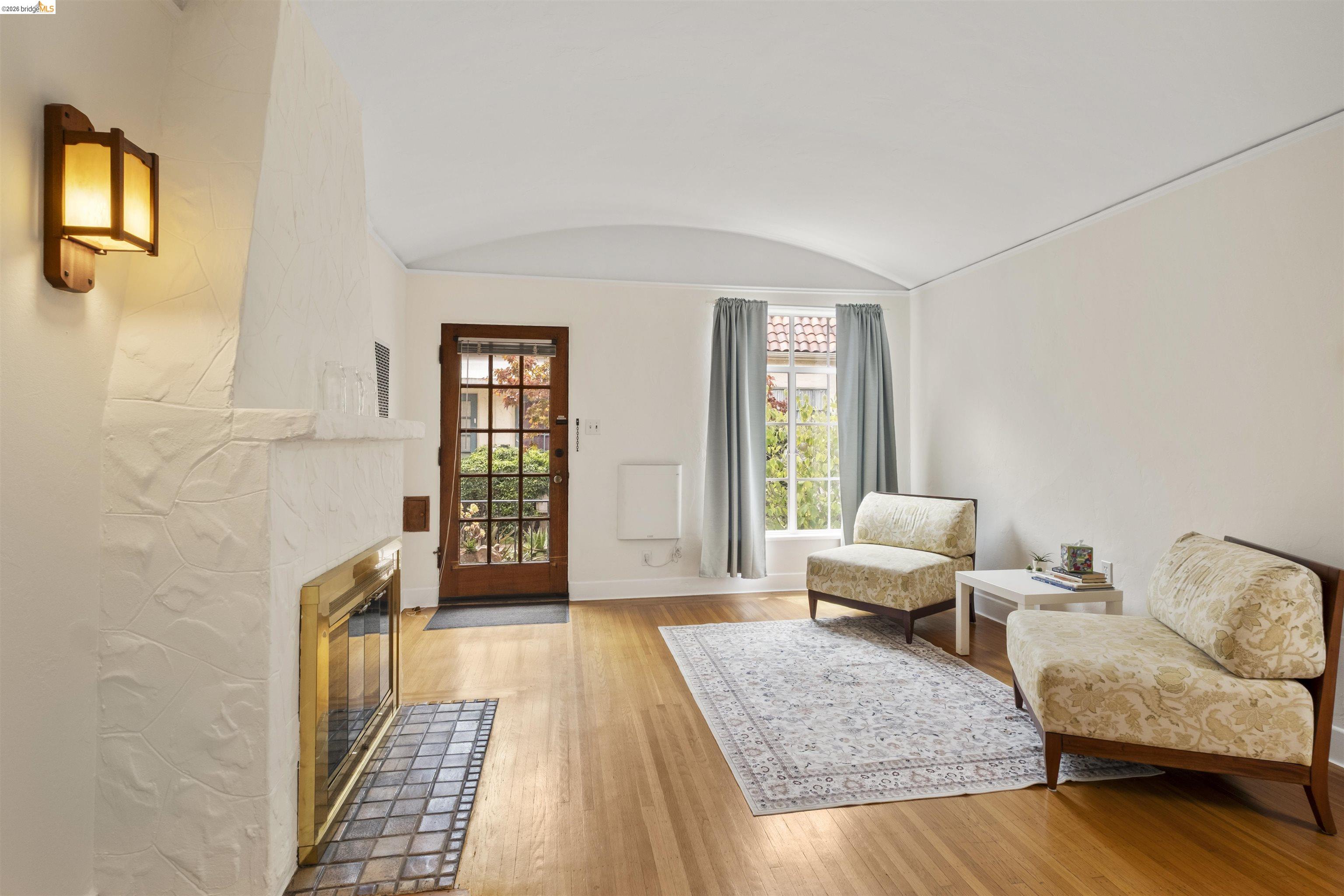 2444 Virginia Street, Unit B Berkeley, CA 94709 - Photo 14 of 39 Living area featuring wood-finish flooring, a textured white fireplace with a brass-trimmed firebox and tile hearth, a glass-paned entry door, and a built-in wall sconce