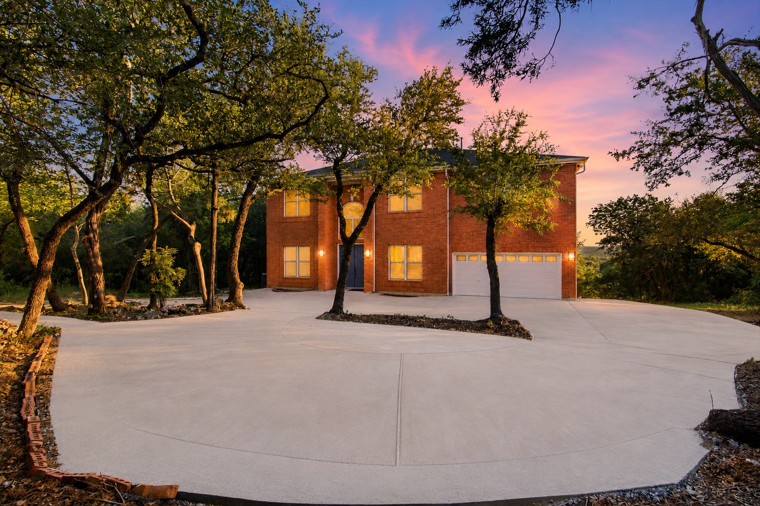 8204 Texas Plume Road Austin, TX 78759 - Photo 1 of 40 A classic brick exterior with a broad concrete driveway is framed by mature trees, all set against a vibrant sunset.