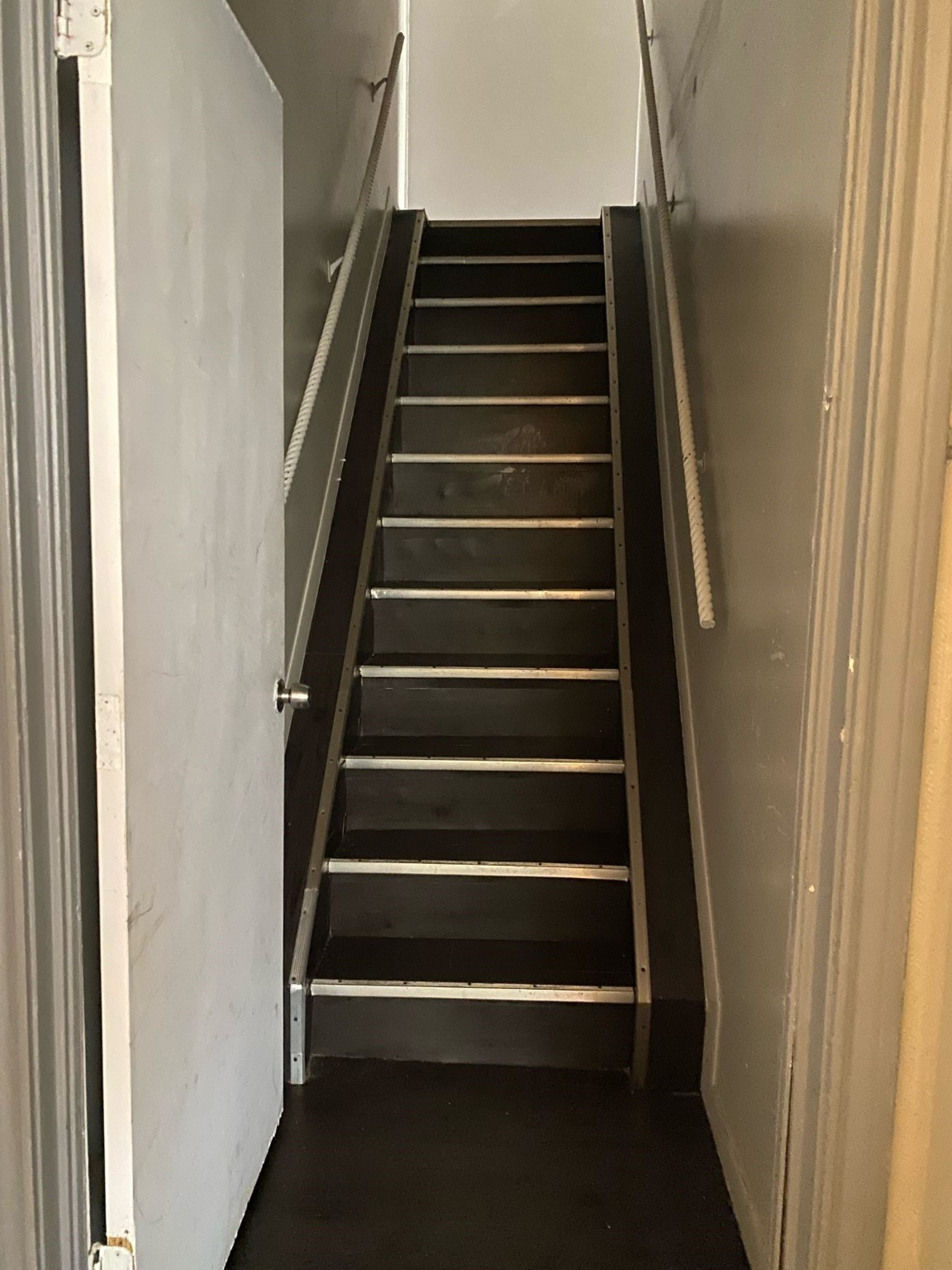 800 Travis Street, Unit ATTIC Columbus, TX 78934 - Photo 2 of 6 The photo shows a narrow staircase with dark steps and metal edges, flanked by plain walls and featuring simple handrails on both sides.