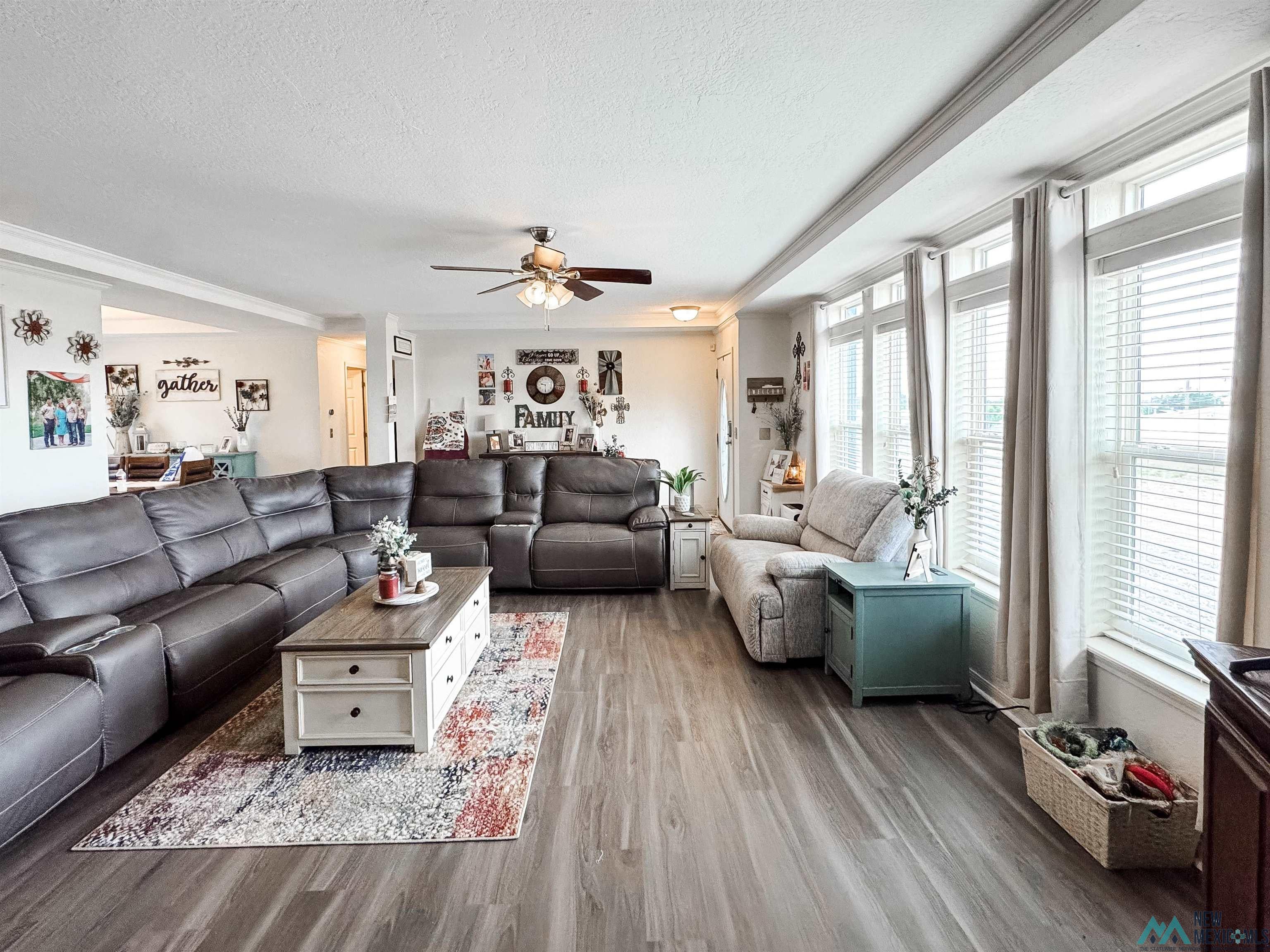 Spacious living area featuring wood-finish flooring, abundant natural light from multiple windows, crown molding details, and a ceiling fan