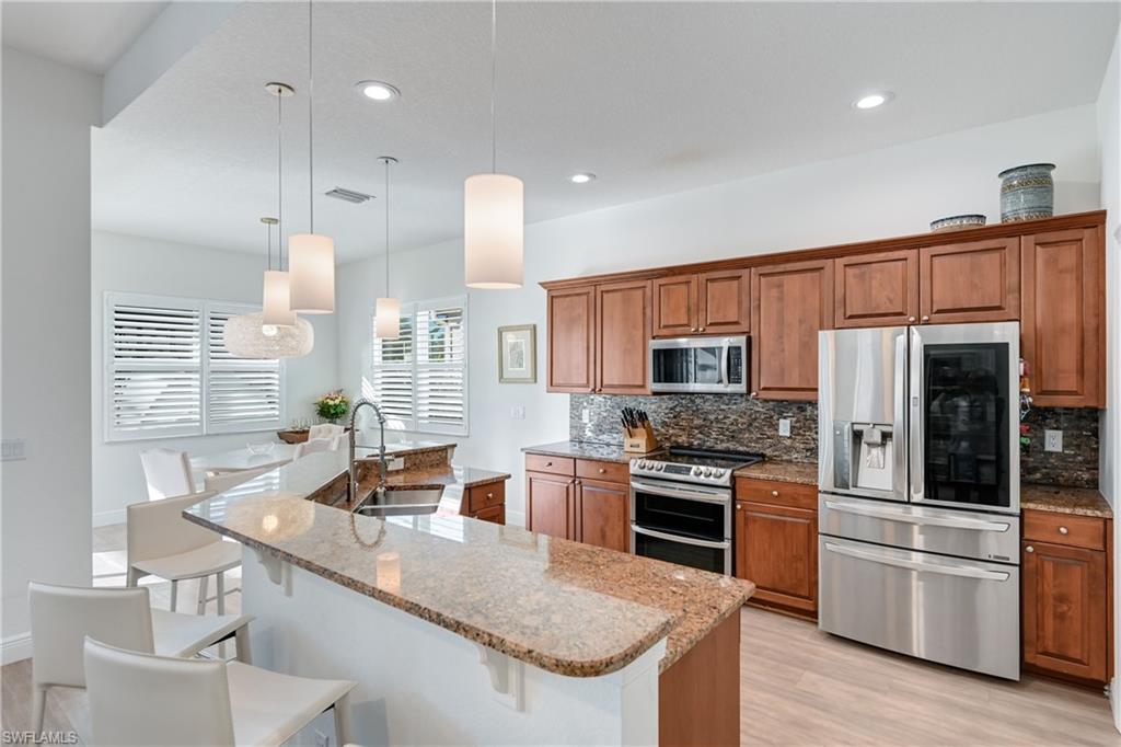 9121 Quartz Lane Naples, FL 34120 - Photo 6 of 36 Kitchen featuring stainless steel appliances, brown cabinets, light stone countertops, decorative light fixtures, and decorative backsplash