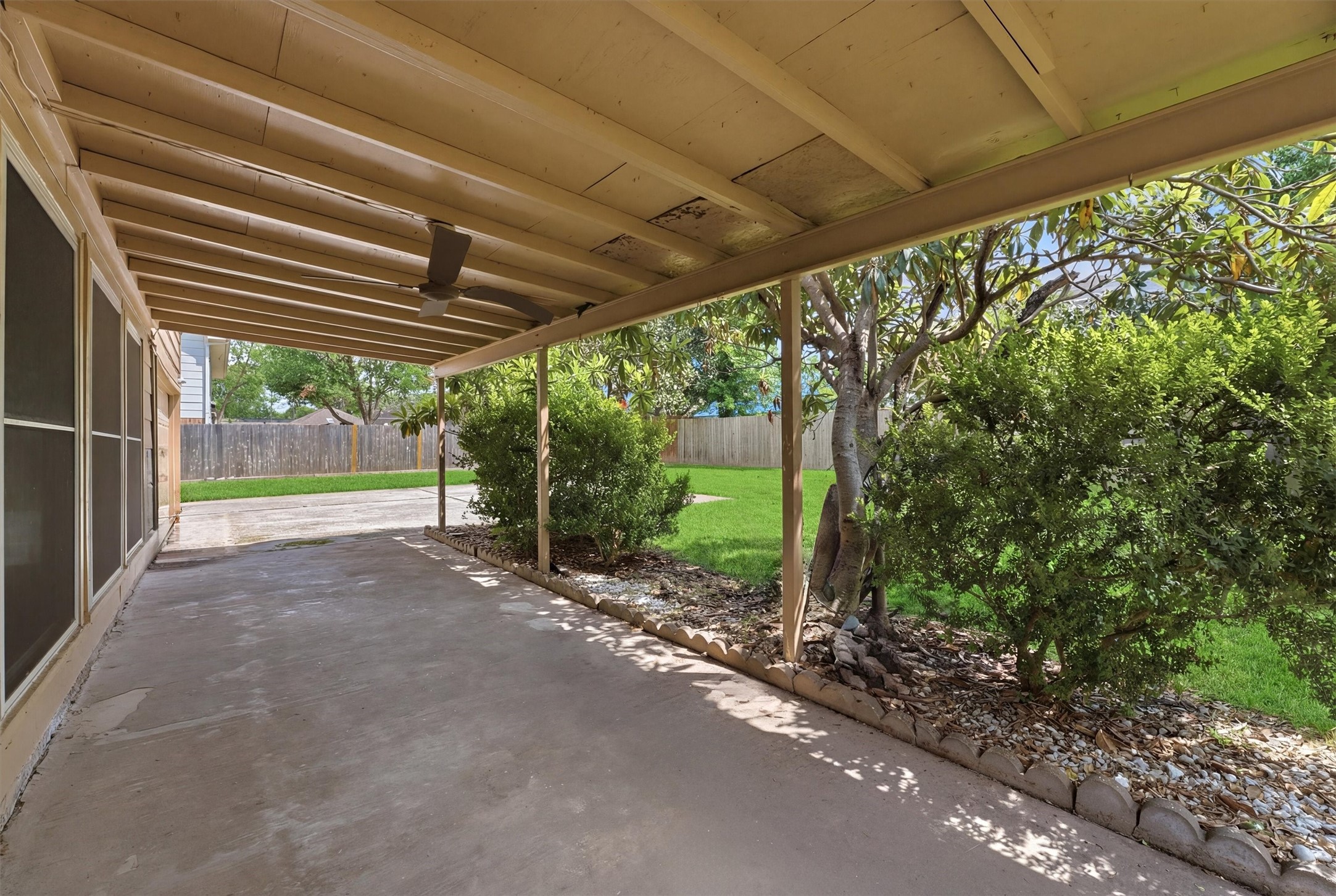 8115 Theisswood Road Spring, TX 77379 - Photo 30 of 41 This photo showcases a covered patio area with a concrete floor, perfect for outdoor relaxation. It opens up to a lush, fenced backyard with trees and shrubs, offering privacy and a serene atmosphere.