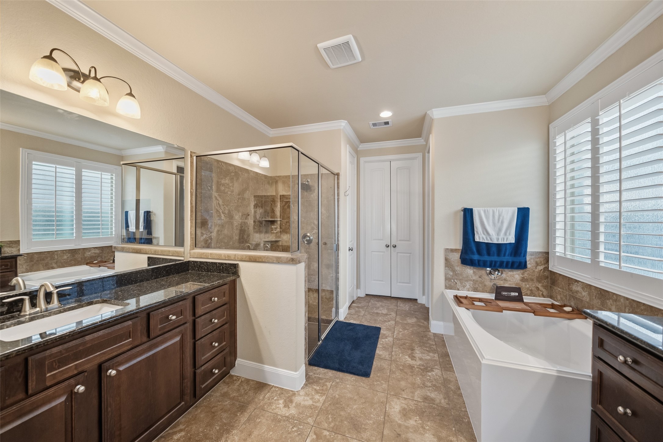 4747 Abercorn Street Rosharon, TX 77583 - Photo 21 of 46 This primary bathroom features a spacious design with a glass-enclosed shower, a luxurious soaking tub, and dual sinks with dark wood cabinetry. It has elegant tile flooring, ample natural light from large windows with shutters, and modern fixtures.