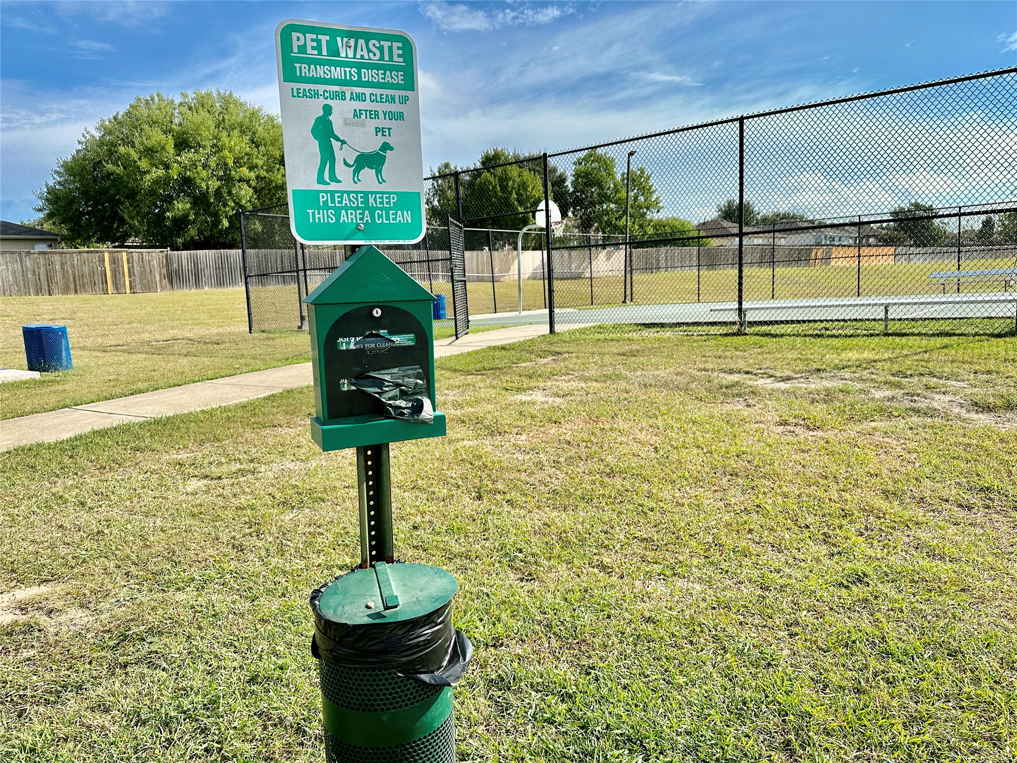 207 Dragon Ridge Road Buda, TX 78610 - Photo 33 of 38 Grassy outdoor area featuring a pet waste station with an integrated receptacle, a paved walkway, and a black chain-link fence enclosing a recreational court