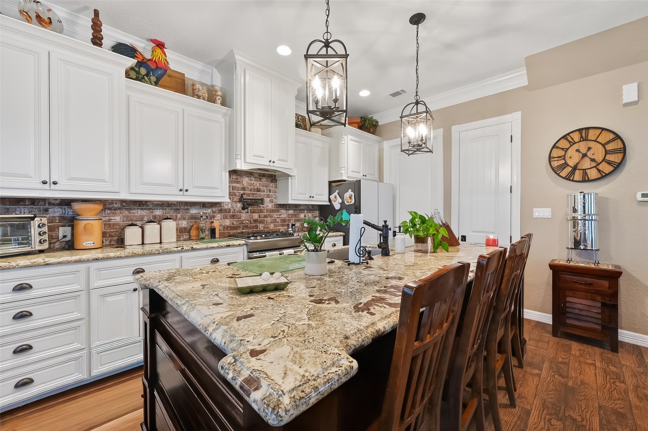 912 County Road 340 Angleton, TX 77515 - Photo 13 of 50 This kitchen features elegant granite countertops, a large island with bar seating, and stylish pendant lighting. White cabinetry contrasts with a brick backsplash, and the space includes modern appliances and warm wood flooring, creating a cozy, inviting atmosphere.