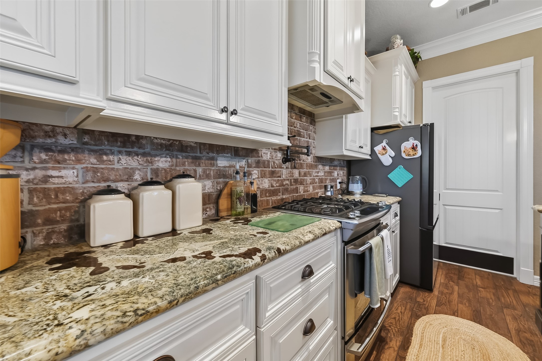912 County Road 340 Angleton, TX 77515 - Photo 14 of 50 This kitchen features elegant white cabinetry, a rustic brick backsplash, and granite countertops. It includes modern appliances, a gas stove, and has wood flooring for a warm, inviting feel.