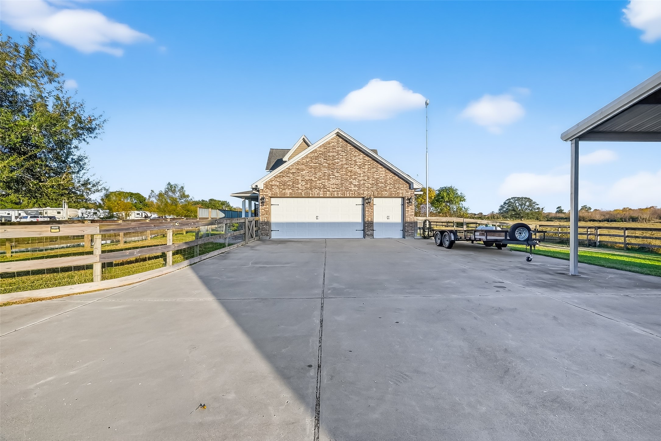 912 County Road 340 Angleton, TX 77515 - Photo 31 of 50 This photo shows a spacious driveway leading to a brick garage with three doors, set in a rural area. There's a wooden fence to the left and a carport on the right, with open land in the background.