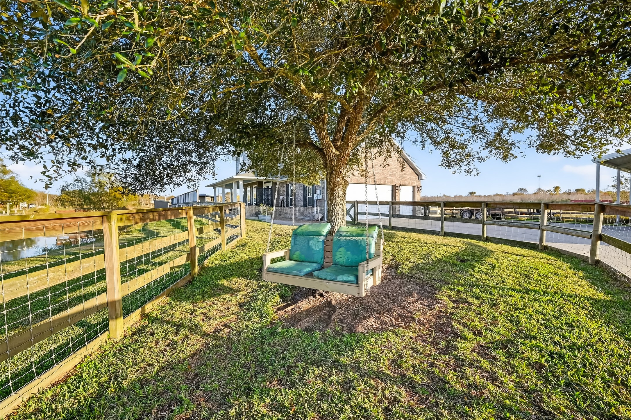912 County Road 340 Angleton, TX 77515 - Photo 32 of 50 Charming outdoor space featuring a cozy swing under a tree, surrounded by a well-maintained lawn and wooden fencing. A picturesque pond and a modern barn-style building are visible in the background, creating a serene, inviting atmosphere.