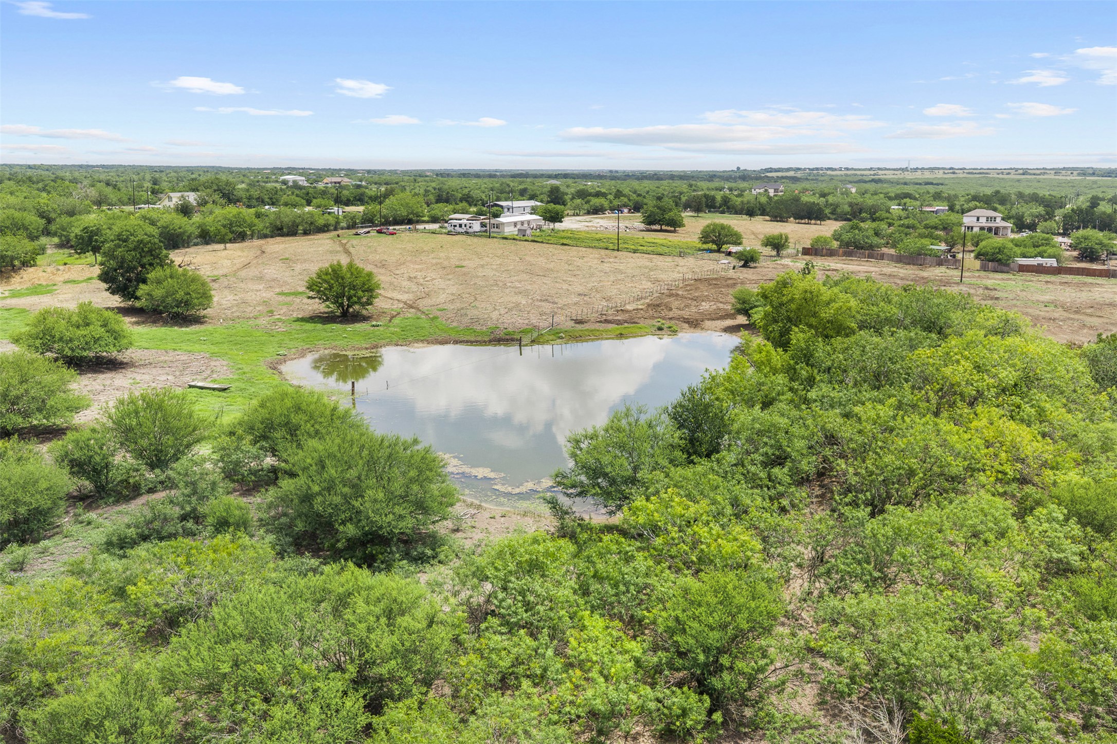 Discover a tranquil rural landscape with a shimmering pond reflecting the sky, surrounded by verdant trees and open fields.