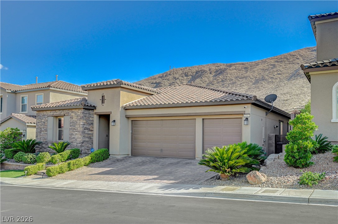 3943 Reyes Avenue Las Vegas, NV 89141 - Photo 2 of 95 A desert-modern home with a two-car garage features stone accents and a tiled roof, set against a mountain backdrop.