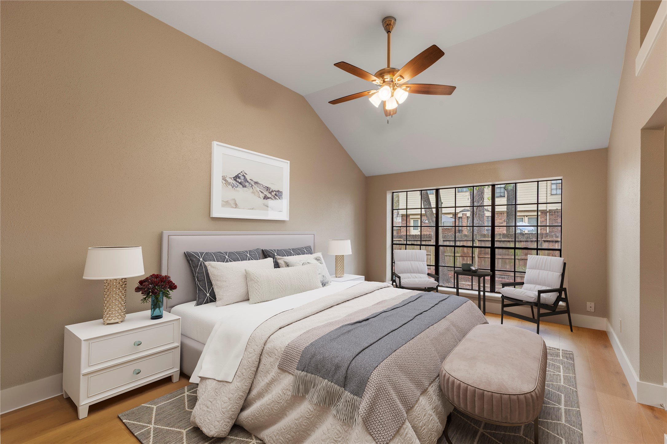 10 White Fawn Drive Spring, TX 77381 - Photo 19 of 26 This primary bedroom features a vaulted ceiling with a ceiling fan, a large window providing natural light, and a cozy seating area. It's elegantly furnished with a plush bed, bedside tables, and soft lighting, creating a warm and inviting atmosphere. Virtually staged.