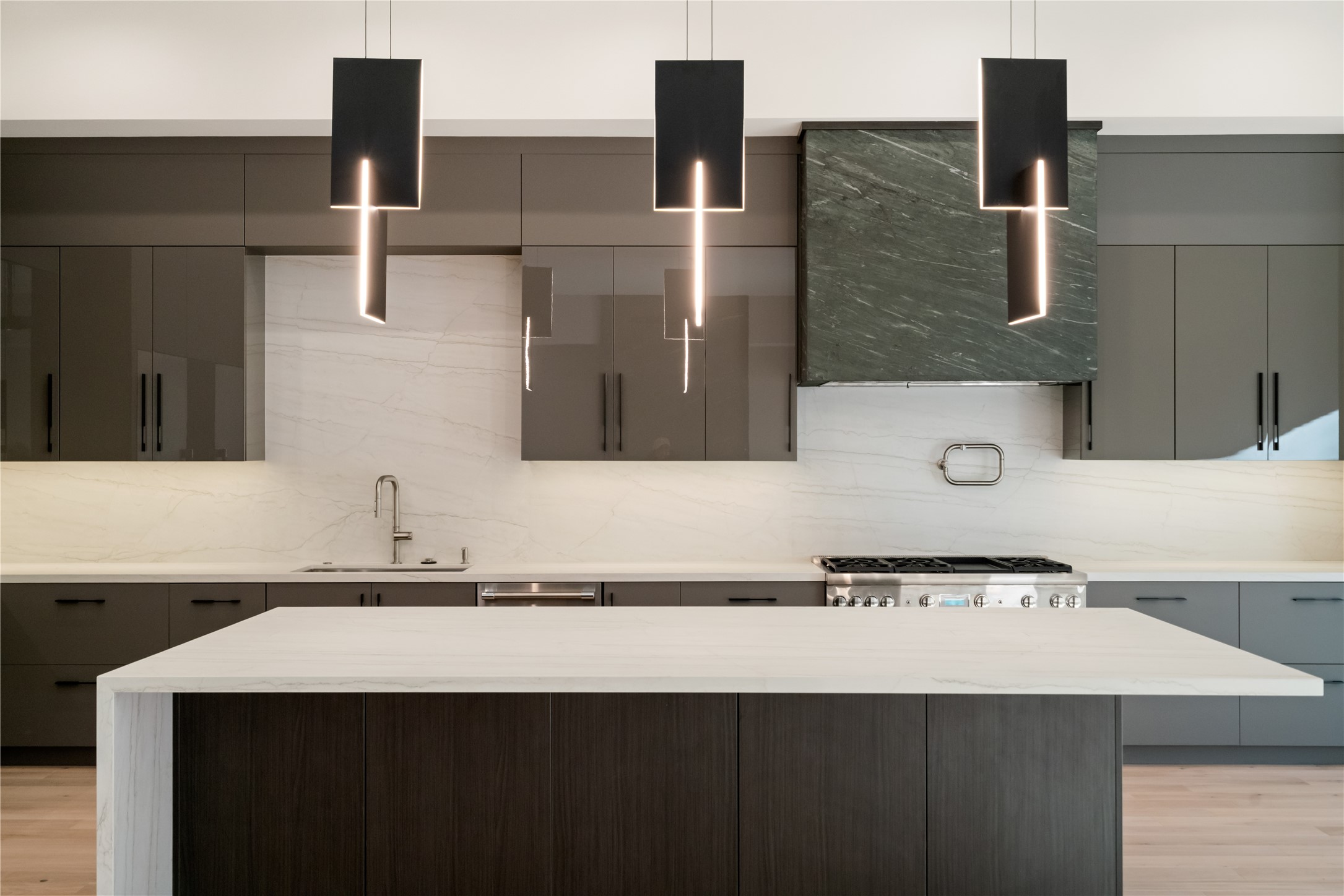 47 Pronghorn Place Spring, TX 77389 - Photo 10 of 44 Sleek modern kitchen featuring dark cabinetry, a large marble island, and contemporary pendant lights. The backsplash and countertop provide a clean, elegant look, with a high-end stove and hood for a sophisticated touch.