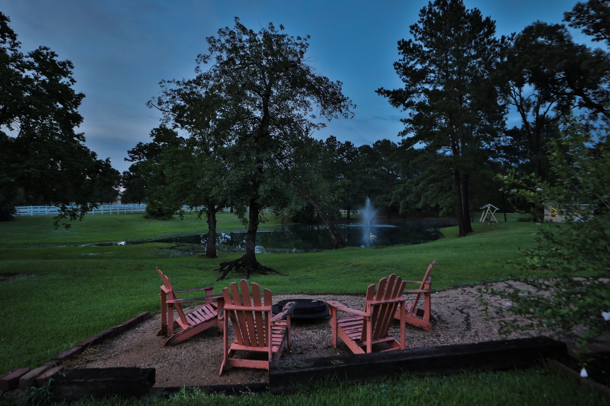 34310 High Point Drive Magnolia, TX 77355 - Photo 42 of 48 This photo showcases a serene property at dusk, featuring a spacious yard with mature trees, a tranquil pond with a fountain, and room for your cozy seating around a fire pit. Perfect place to unwind with your guests!
