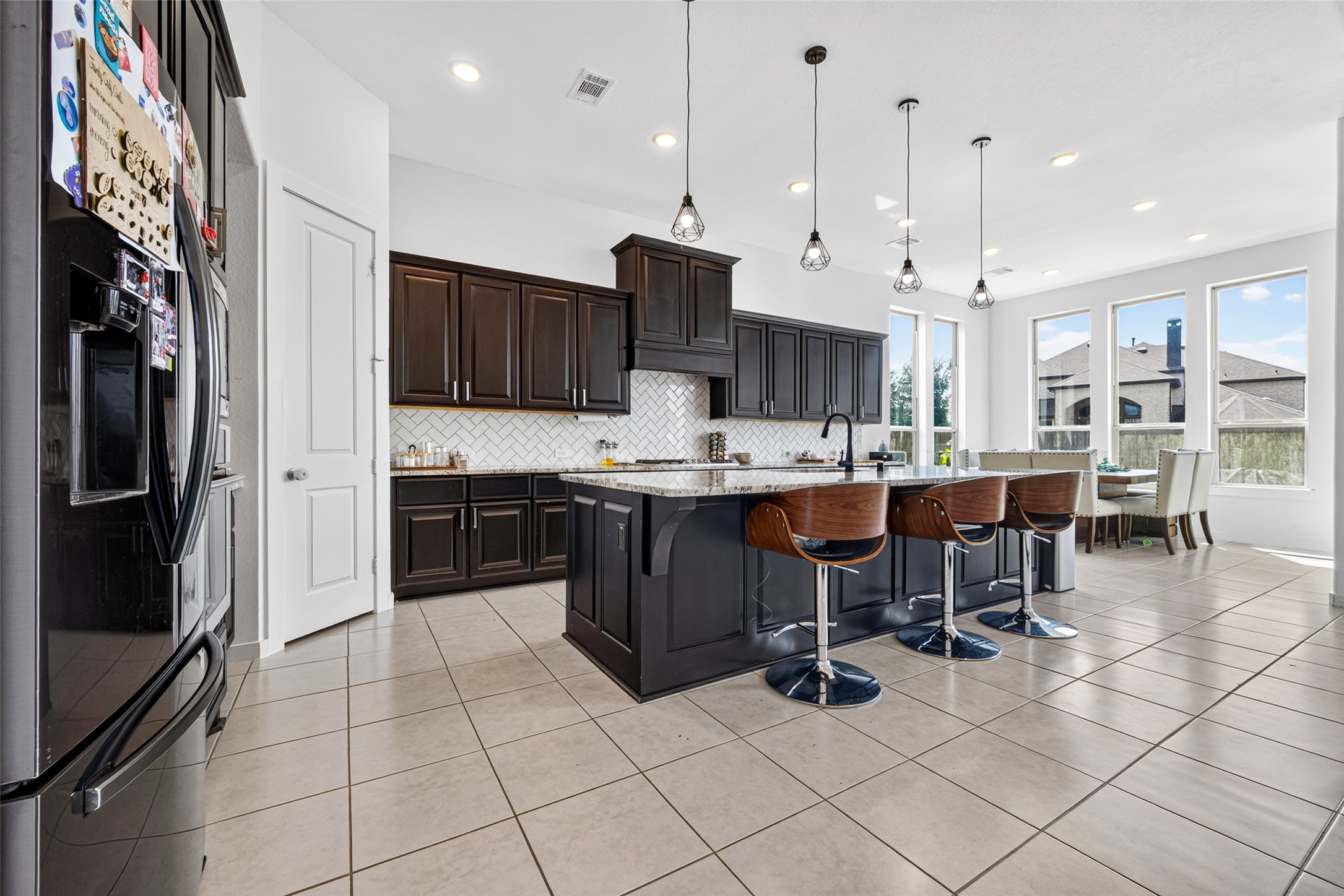 10114 Brush Way Rosharon, TX 77583 - Photo 16 of 50 Stunning modern kitchen featuring dark wood cabinetry, a spacious island with bar seating, and elegant pendant lighting. The large windows fill the space with natural light, enhancing the open layout and providing a welcoming dining area.
