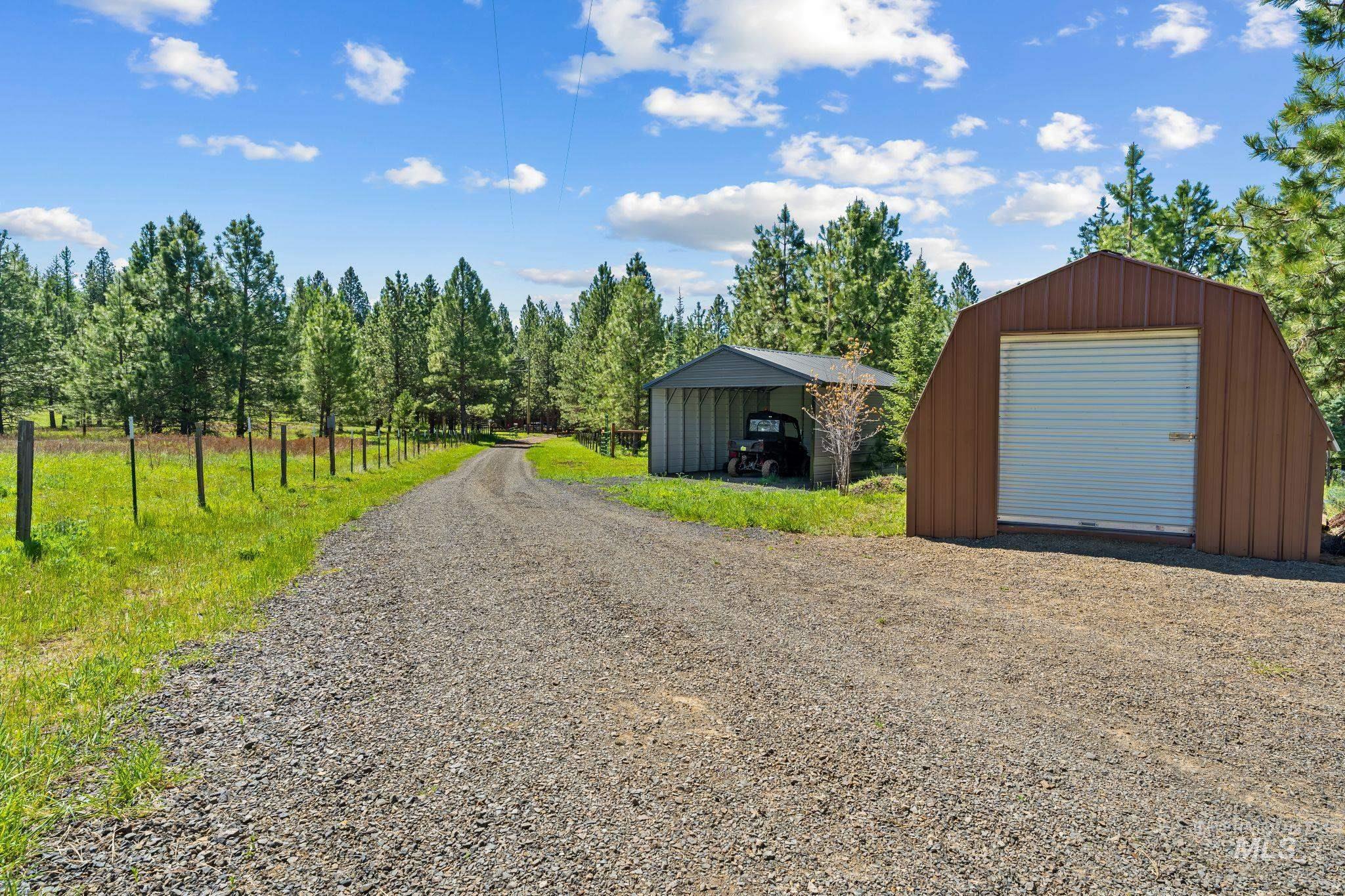507 Bearlea Road Anatone, WA 99401 - Photo 25 of 47 Gravel driveway extending through a wooded property, featuring a brown metal Quonset-style shed with a roll-up door and an open-sided metal carport