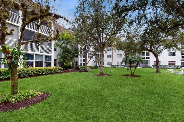 a view of a white house in front of a big yard with plants and large trees