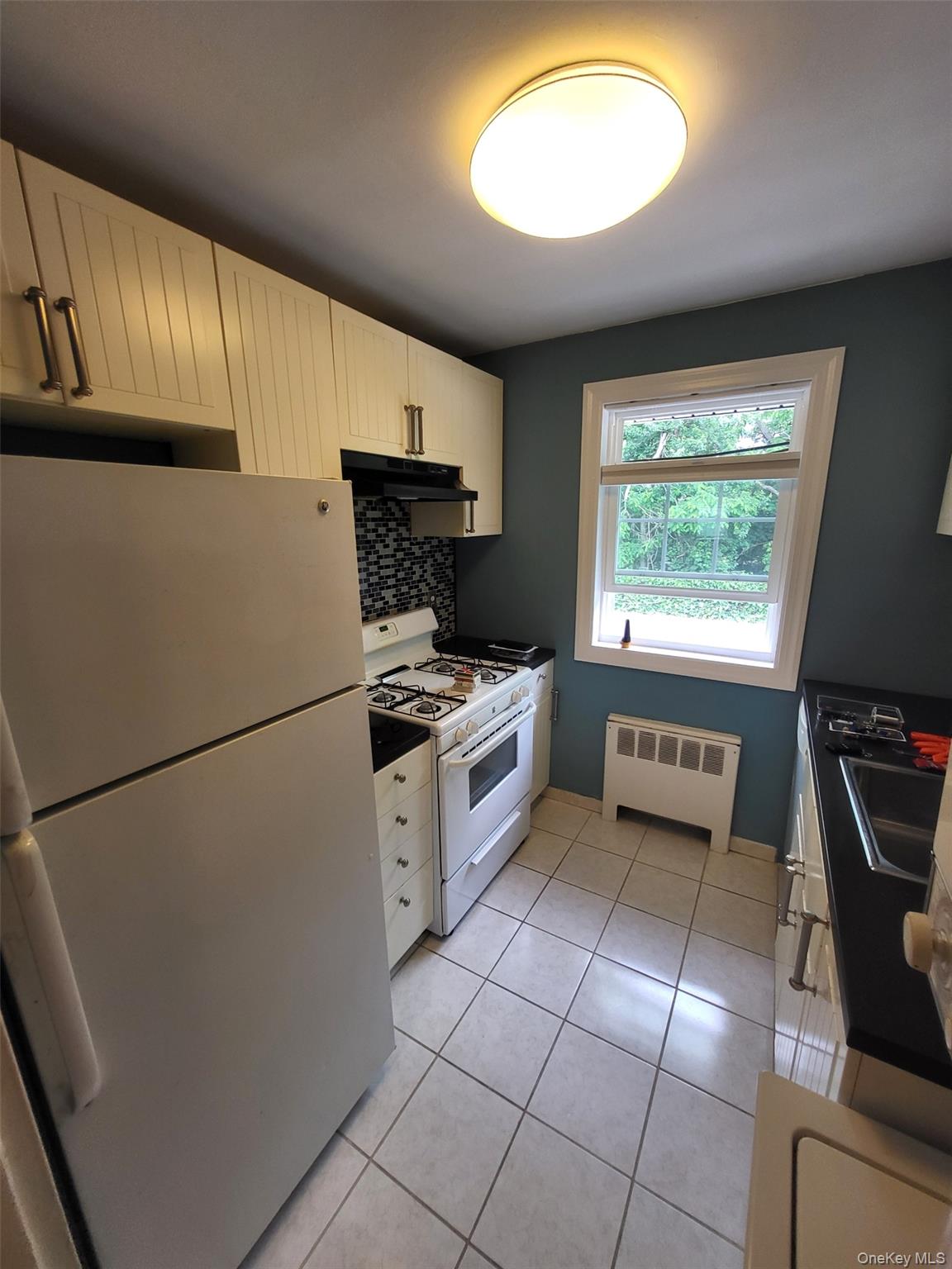 224-23 Kingsbury Avenue, Unit B Queens, NY 11364 - Photo 16 of 22 Kitchen with white appliances, radiator heating unit, under cabinet range hood, tasteful backsplash, and light tile patterned flooring