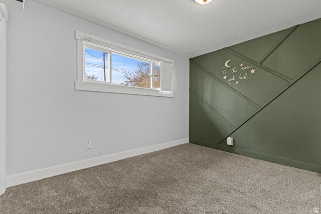 2101 West 3900th Street South Roy, UT 84067 - Photo 16 of 36 Room featuring a decorative accent wall with geometric paneling, natural light from a window, neutral-toned carpeting, white baseboards, and a flush-mount ceiling light