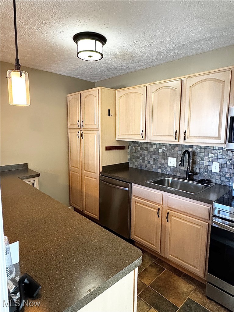 Kitchen with light brown cabinets, dark countertops, stainless steel appliances, backsplash, and a textured ceiling