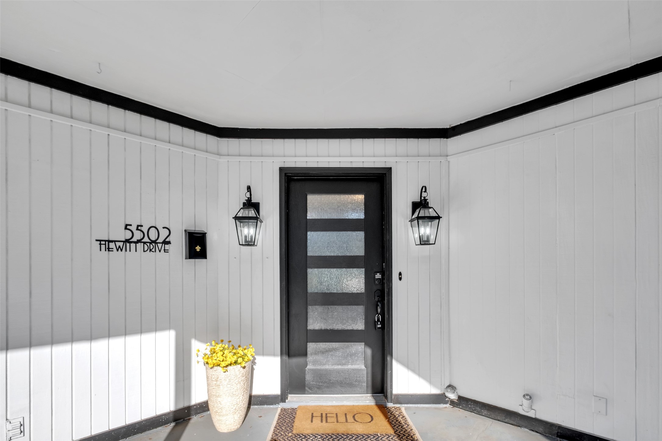 This inviting entrance features a modern black door with frosted glass panels, framed by sleek black lanterns. The white paneling adds a clean, crisp look. The address is elegantly displayed, creating a stylish and welcoming first impression.