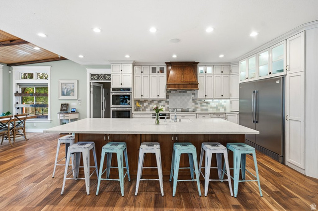 672 Perry Hollow Drive Mapleton, UT 84664 - Photo 26 of 106 Spacious kitchen featuring a large central island with seating, stainless steel appliances, white cabinetry with display uppers, a wood-finish range hood, and wood-finish flooring