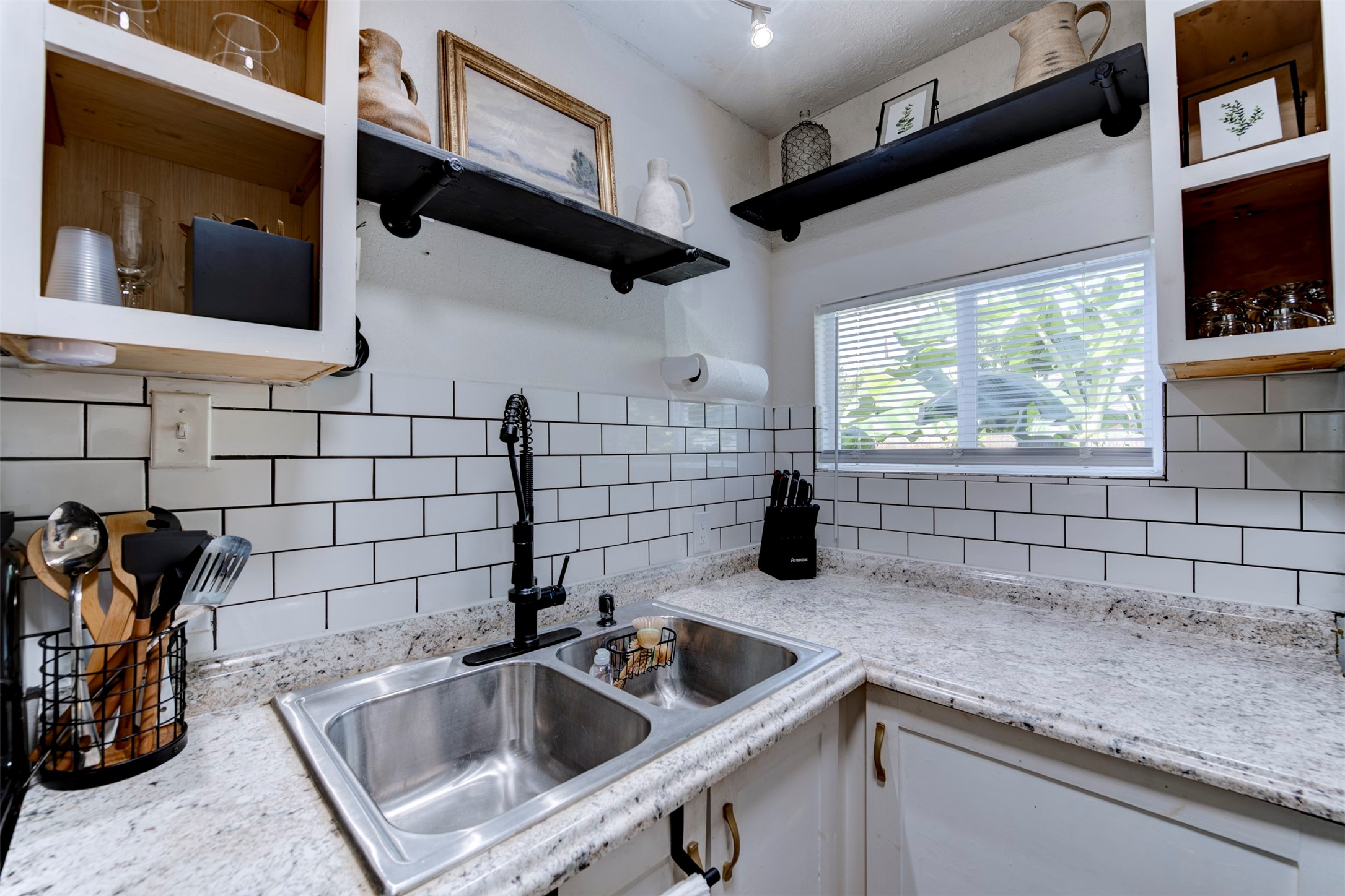 1121 Panama Street Houston, TX 77009 - Photo 12 of 35 Kitchen sink area featuring double-basin stainless steel undermount sink, matte black pull-down spring faucet, granite countertops, white subway tile backsplash with dark grout, track lighting, open wood cabinets with natural pine interiors, and dual black pipe bracket floating shelves.