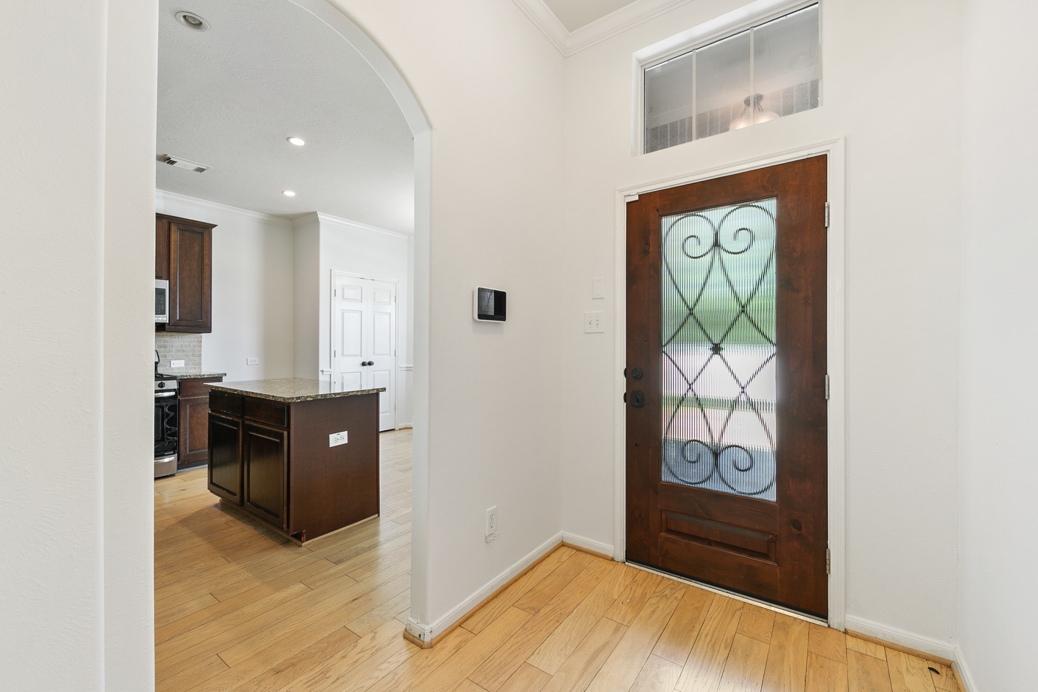 24527 Spring Harbor Drive Spring, TX 77373 - Photo 3 of 22 This photo showcases a welcoming entryway with a decorative wooden door featuring glass insets. The space has light hardwood flooring and leads into a kitchen with dark cabinetry and a central island, offering a bright and inviting entrance.