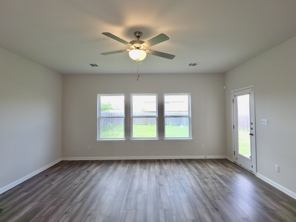 110 Grapefruit Road Hutto, TX 78634 - Photo 2 of 28 Spacious living room featuring wood-finish flooring, light neutral wall paint, three large windows, a ceiling fan with integrated lighting, and a glass-paneled exterior door