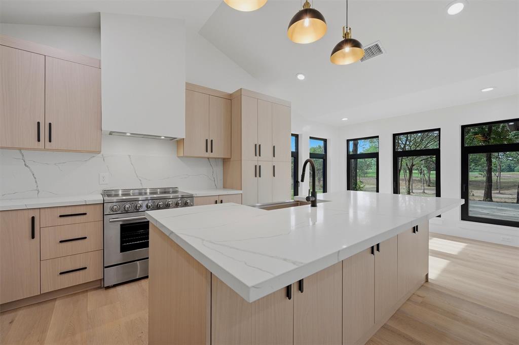 410 Arkansas Road Sadler, TX 76264 - Photo 12 of 40 Modern kitchen featuring a large central island with a quartz countertop, light wood-finish cabinetry, a stainless steel professional-grade range, an integrated range hood, and black frame windows