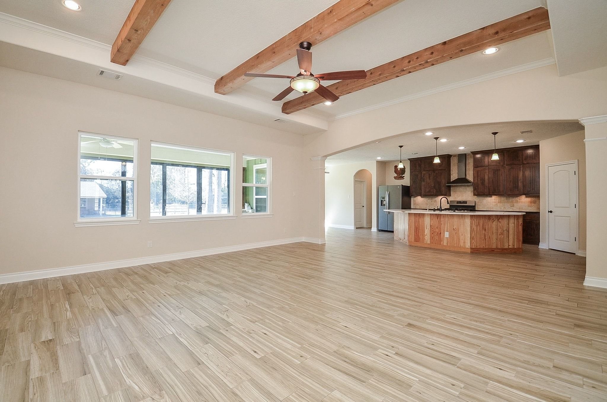 13327 Little Ranch Road Cypress, TX 77429 - Photo 13 of 49 This spacious open-concept living area features wood-like flooring, exposed wooden ceiling beams, and ample natural light from large windows. It seamlessly connects to a modern kitchen with a large island, pendant lighting, and dark wood cabinetry.