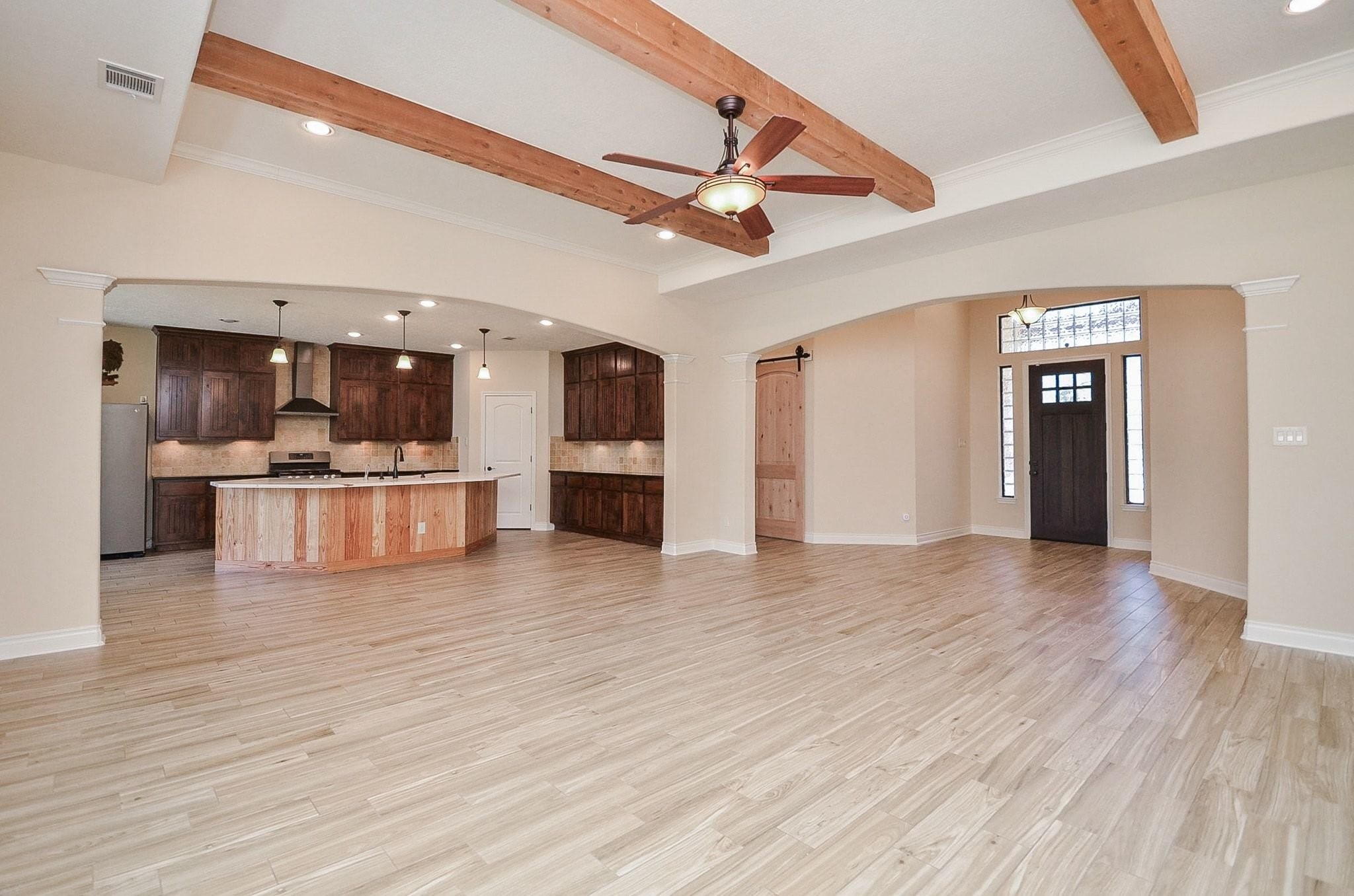 13327 Little Ranch Road Cypress, TX 77429 - Photo 15 of 49 This spacious open-concept living area features light wood flooring, exposed wooden beams, and a ceiling fan. The kitchen boasts dark wood cabinetry, a large island with a natural wood finish, and modern pendant lighting. A welcoming entryway with a dark wooden door and sidelights completes the inviting atmosphere.